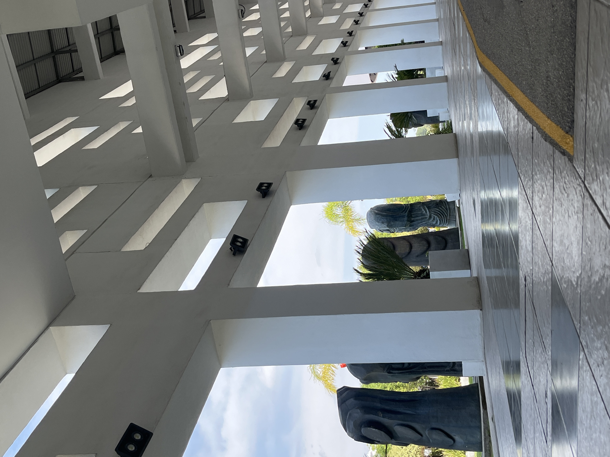 The image shows a building exterior in Unknown, Unknown. The architecture features repetitive white structural elements casting linear shadows on the ceiling. Wall-mounted lights are visible. Outside the structure are plants and statues. A tiled walkway with a yellow stripe borders the building.