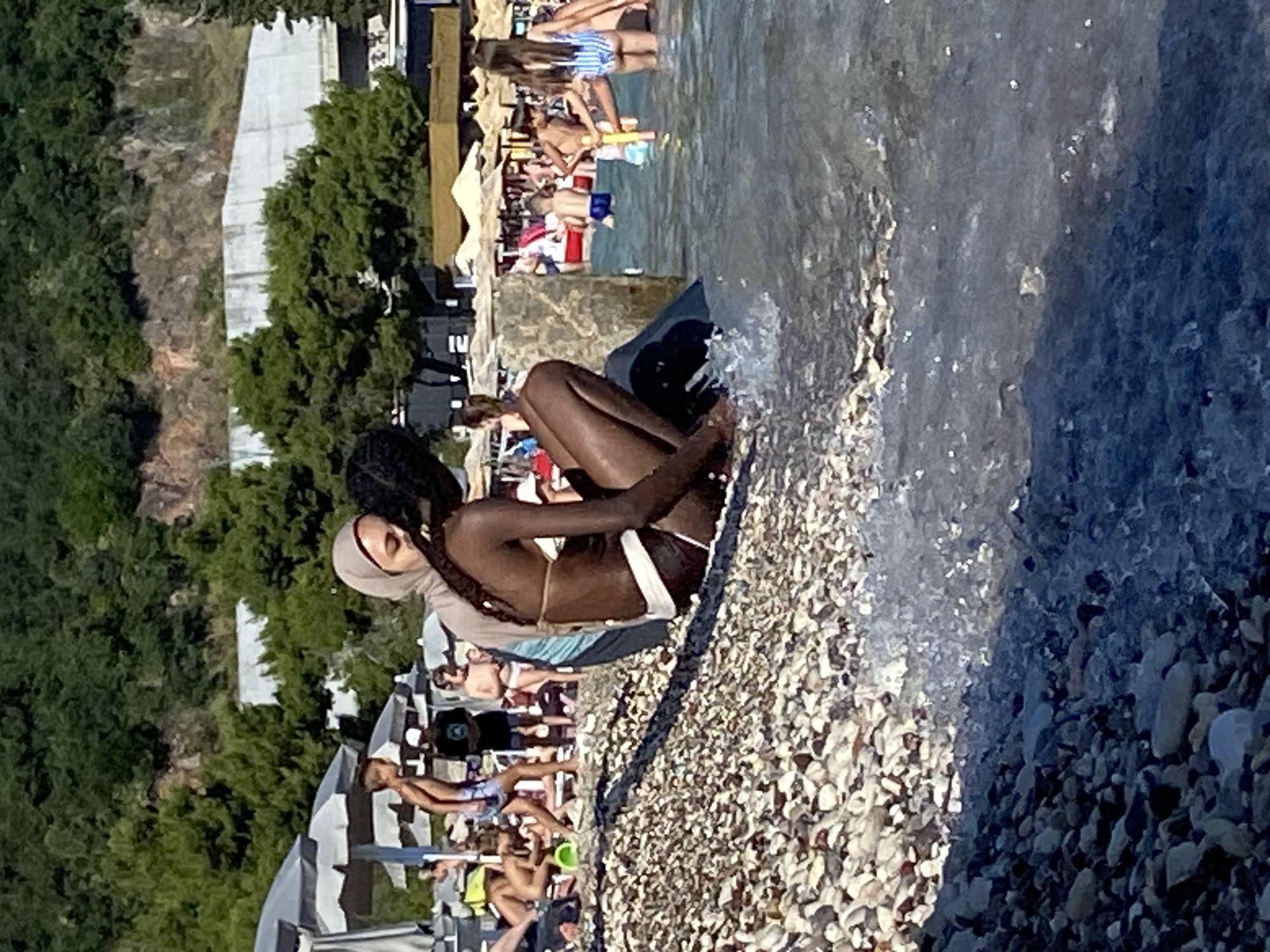 The image depicts a crowded beach in Budva, Montenegro. In the foreground, two individuals are seated on a pebble beach near the water's edge. One person, wearing a hijab and a bikini top, is embracing another person, who is wearing a bikini. The person being embraced is darker-skinned and appears to be facing away from the viewer. Small waves are lapping at the shore, creating white foam.
In the background, numerous beachgoers are visible. Several are lying on sunbeds under umbrellas, while others are walking or standing. Children are playing in the shallow water. The beach is backed by a densely forested hillside, with a concrete structure visible partway up the slope. Beach umbrellas, lounge chairs, and some low buildings with awnings are also present. The overall scene suggests a sunny day at a popular tourist destination.