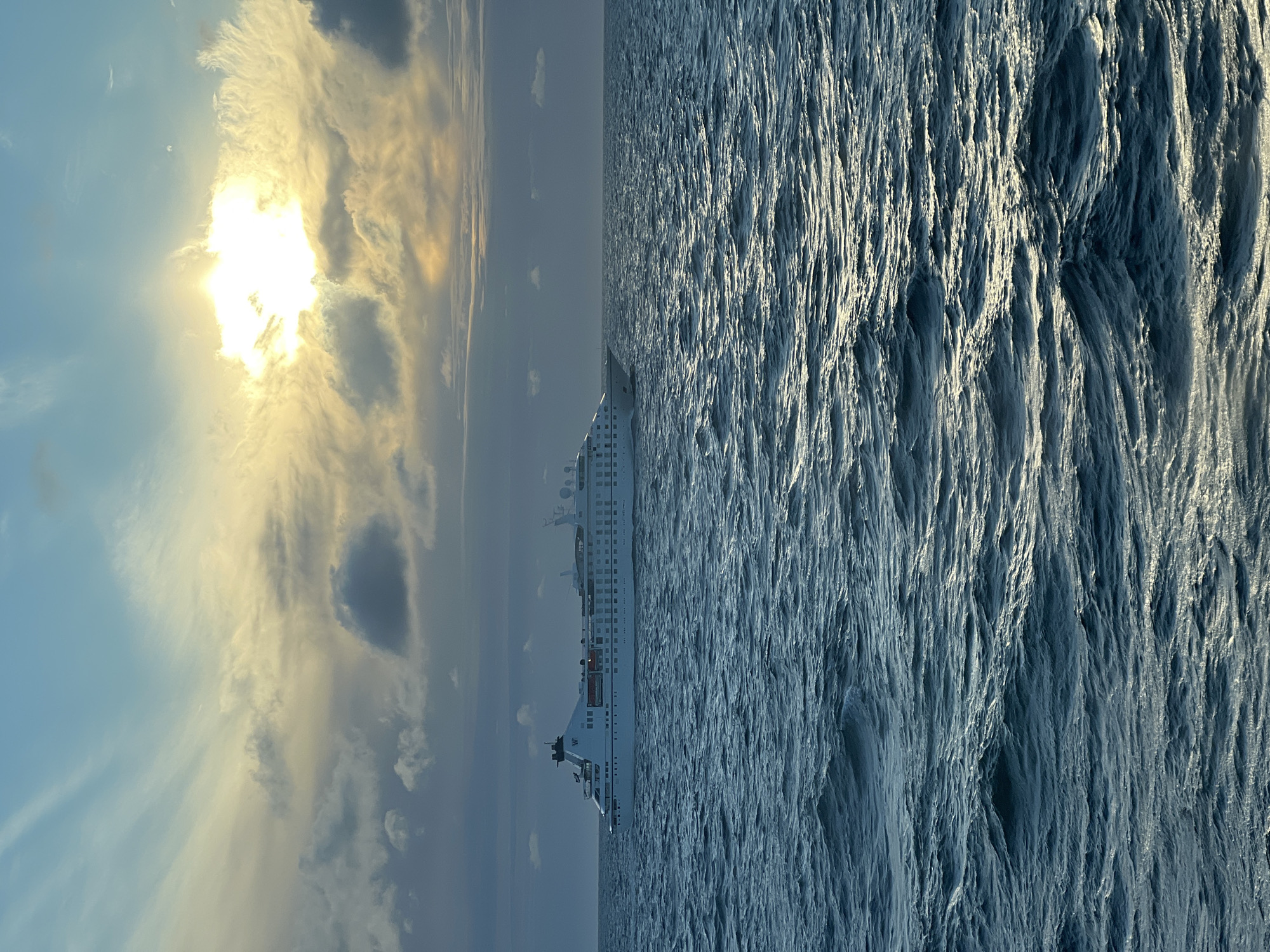 A wide-angle, high-angle shot shows a cruise ship sailing on the ocean under a partly cloudy sky. The sun shines brightly through a break in the clouds, casting a warm glow on the water. The ship, a white multi-deck vessel, is positioned in the middle left of the frame, sailing from left to right. The ocean is a deep blue, with whitecaps on the waves. The sky is a mix of blues, grays, and yellows, with streaks of clouds. The location is unknown, Poland.