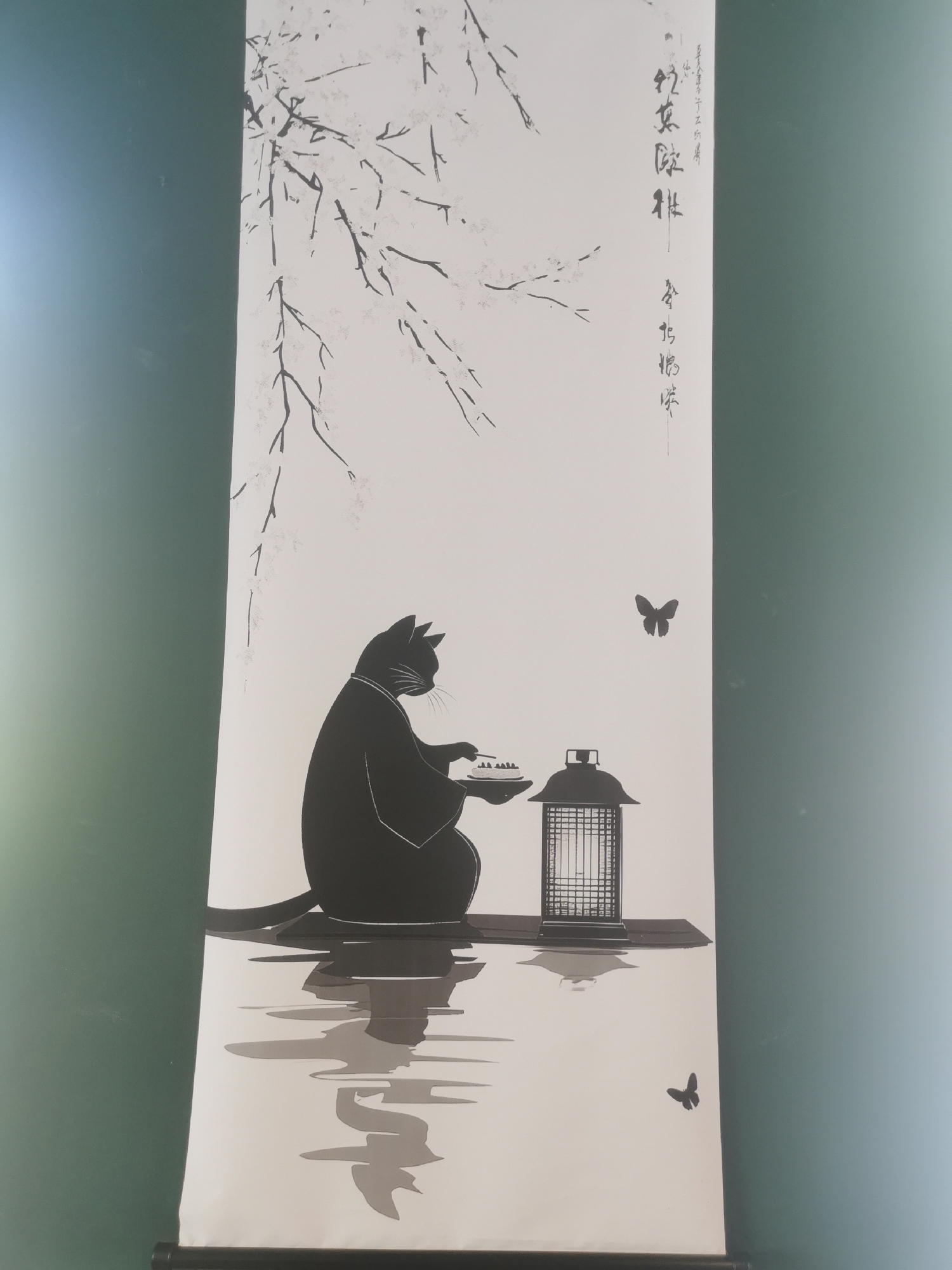 The image displays a vertical artwork, likely a scroll or poster, featuring a minimalist black and white illustration, hanging against a plain teal-green wall.
The central subject of the artwork is a black cat, depicted in profile and silhouette, seated on a platform that appears to float on water. The cat wears a traditional-style robe or kimono and has its paws resting near a small dish containing several small, white objects, suggesting a quiet activity such as a tea ceremony or a meal. Beside the cat is a traditional East Asian lantern, also rendered in silhouette. The cat, platform, and lantern are reflected in the calm water below, depicted with stylized ripples.
In the upper left portion of the scroll, delicate, leafless tree branches, possibly cherry blossom or willow, extend downwards. Two small black butterflies are seen in the white space: one above and to the right of the lantern, and another near the bottom right.
The artistic style is reminiscent of traditional Japanese ink painting (sumi-e) or woodblock prints, characterized by strong black forms against a white background, creating a serene and contemplative mood. Vertical calligraphic text, likely Japanese or Chinese, is visible on the upper right side of the scroll. The overall scene evokes a sense of tranquility and peaceful introspection. The bottom edge of the scroll is secured by a visible black bar.