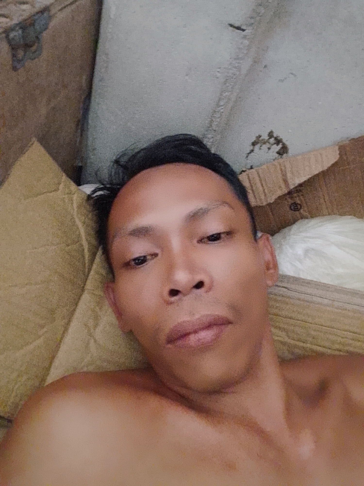 A selfie taken by a Southeast Asian male. He has dark, short hair and is lying down, shirtless, amidst cardboard boxes and a beige pillow. A white object, possibly a crumpled cloth, is visible behind him. The background appears to be an unfinished or cluttered interior. Location: possibly Bukit Ketok, Indonesia.
