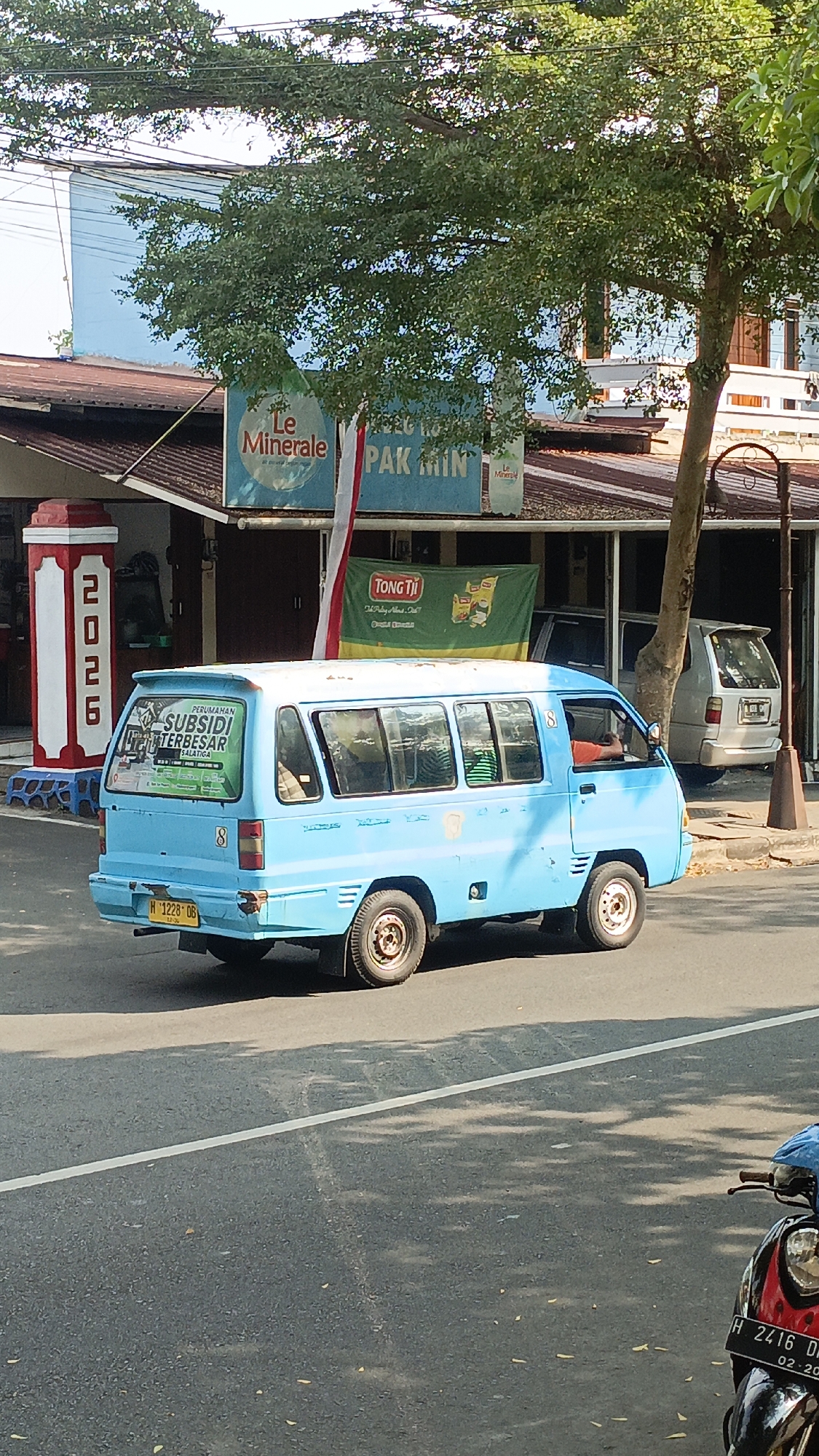 An eye-level, medium shot captures a vibrant street scene in Salatiga, Indonesia, bathed in bright daylight. In the foreground, a section of a paved road with a white line is visible, partially shaded by dappled sunlight filtering through the leaves of a large tree. To the right, the handlebars and headlight of a parked motorcycle with license plate H 2416 DK are prominent.
Dominating the mid-ground is a bright blue minibus with a white roof, seen from its rear-left quarter as it travels on the road. The minibus has several tinted windows and a prominent green sticker on its rear window that reads "PERUMAHAN SUBSIDI TERBESAR SALATIGA". Its license plate is H 1228 DB. The minibus has a spoiler on its roof.
Behind the minibus, a row of buildings with dark red tiled roofs lines the street. A tall, white pillar with red accents, displaying the numbers "2026" vertically, stands on the left side of the frame. Two signs are visible above the buildings: one features the "Le Minerale" logo and text, and another green banner displays "TONG TJI" with an image of tea boxes. A white car is parked to the right of the minibus, in front of one of the buildings. The environment appears to be an urban street, possibly a commercial area, with trees providing shade.