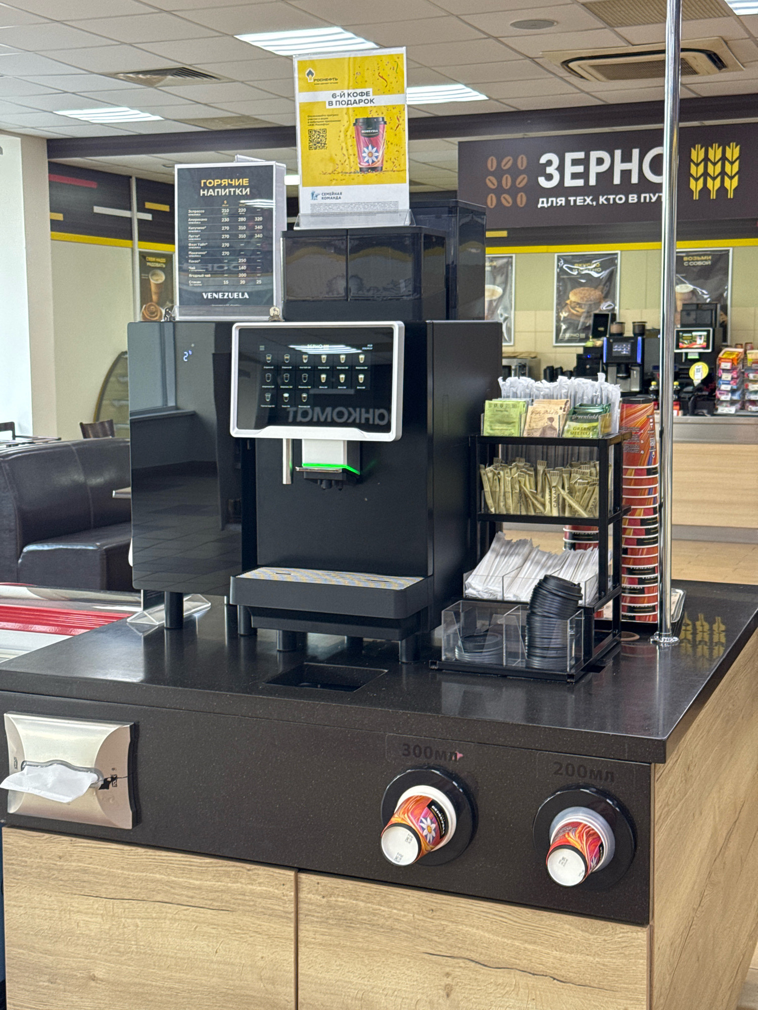 The image shows a coffee machine and a display of coffee drinks in a service station in Novoselitsy, Russia. The coffee machine is black with a digital screen displaying drink options. To the left of the machine is a menu board for hot drinks with prices listed in Russian rubles. To the right, there is a rack with sugar packets, stirrers, and cups. In the background, there are shelves stocked with various goods, and a large sign that reads "ЗЕРНО" which translates to "GRAIN". Two cups are visible on the counter below the machine, one labeled "300мл" (300ml) and the other "200мл" (200ml). The overall environment appears to be indoors, likely a retail or convenience store setting within a gas station. There are no people visible in the image.
