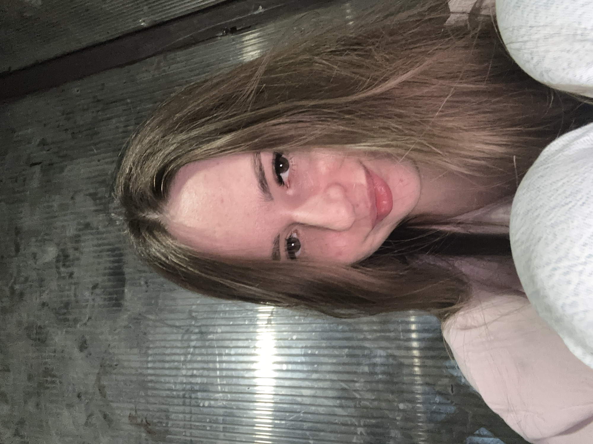 A close-up, rotated selfie shows a young person, likely female, looking directly at the camera with a soft smile. The image is rotated 90 degrees clockwise from a standard portrait orientation. The subject has long, light brown hair framing their face, with their forehead, eyes, nose, and lips clearly visible. Makeup, possibly mascara or eyeliner, is visible around their left eye. Their lips are slightly parted, hinting at teeth. A light-colored, textured garment, possibly a blanket or soft clothing, is visible on the right side of the frame. The background is dark and appears to be a metallic or corrugated surface with prominent horizontal ridges, suggesting an enclosed or utilitarian space. A bright vertical light reflection is visible on the left side of the background. No text is visible. The person appears to be in a relaxed and pleasant mood.