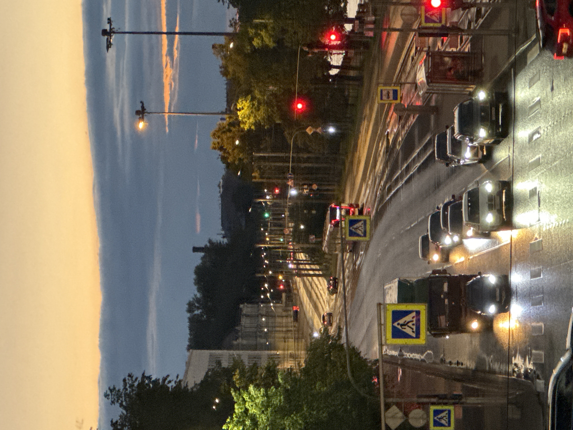 The image depicts a street scene in Kudrovo, Russia, during dusk. The sky is transitioning from a pale yellow to a muted blue, with wisps of orange clouds visible near the horizon. A prominent feature is a wide, wet road with multiple lanes, indicated by the reflections of headlights and taillights on the asphalt. Several vehicles are present, including cars, a truck, and what appears to be a bus or tram in the distance. Traffic lights are visible, some showing red, suggesting traffic is stopped or slowed. Pedestrian crossing signs with yellow backgrounds and blue symbols are also present. Streetlights illuminate sections of the road and sidewalks. Trees line the right side of the road, and buildings are visible in the background, some with illuminated windows. The overall atmosphere is one of a busy urban environment at the end of the day.