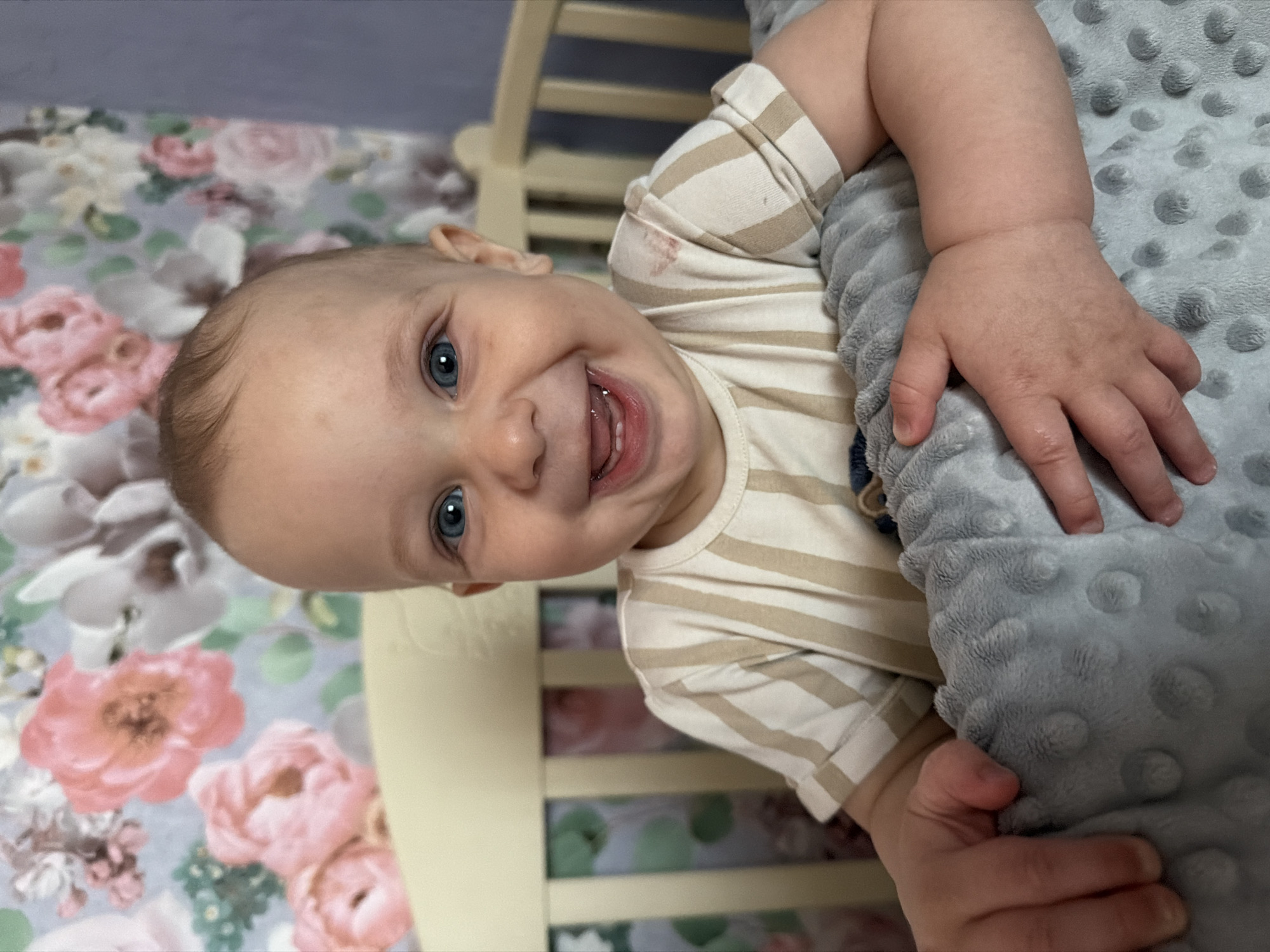 A baby is in a crib, the image is rotated 90 degrees counter-clockwise. The baby is smiling broadly, with a wide open mouth showing teeth and tongue. The baby has blue eyes and fair skin with a few wisps of light brown hair. The baby is wearing a striped shirt with beige and white stripes. The baby's left arm is bent and resting on a gray, textured blanket. The crib has light-colored wooden slats visible in the background. The crib bedding has a floral pattern with pink and white flowers. The baby appears happy and engaged, looking directly at the camera. The lighting suggests it is daytime. The context provided indicates the location is Kryvyi Rih, Ukraine.