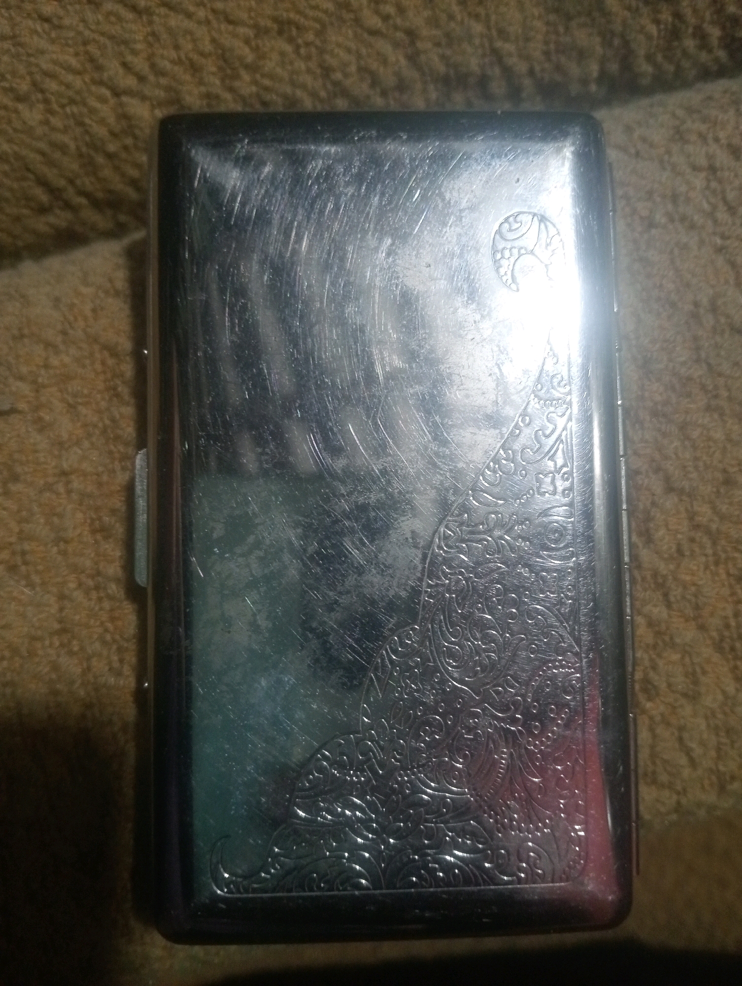 This is a top-down, medium shot of a silver cigarette case with an ornate, engraved design, resting on a brown, textured surface. The cigarette case is rectangular with rounded corners and shows visible scratches and scuff marks on its surface, indicating wear. The engraving features intricate floral and scroll patterns, with a circular emblem near the top right corner. The background is a soft, brown, fabric-like material. There are no people visible in the image. The scene is indoors, with artificial lighting causing reflections and glare on the metal surface of the cigarette case. The location is not specified in the image.