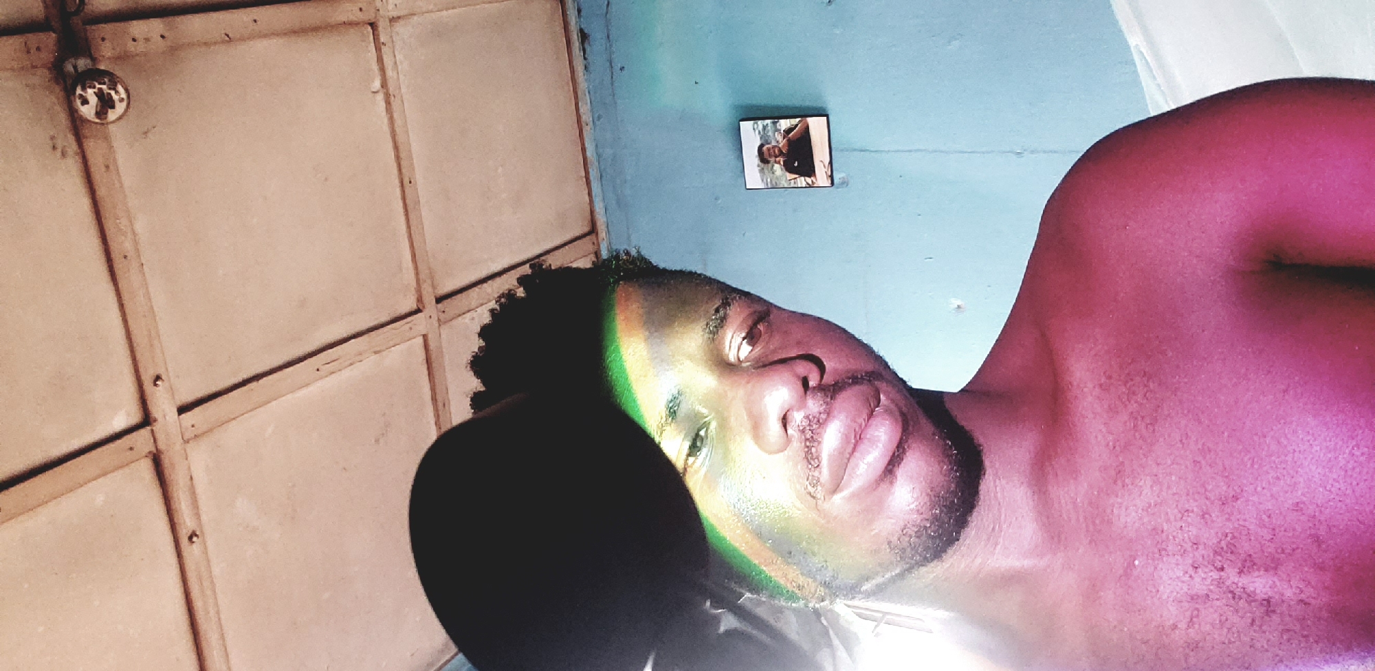 This is a portrait of a young man indoors, likely in Narayi, Nigeria. The man is the primary subject, positioned in the foreground and looking directly at the camera. His face is partially obscured by a shadow cast from above, and illuminated by a green and yellow light that creates a distinct stripe across his forehead and cheek. He has short, curly dark hair and a beard. His skin tone is dark, and the lighting also gives his exposed chest and shoulders a reddish hue.
The background shows a light blue wall with a framed photograph hanging on it. The photograph depicts another young man sitting at a table. Above the wall, a portion of a ceiling is visible, constructed from square panels separated by wooden strips. A circular light fixture, possibly a bulb holder, is attached to the ceiling. The overall impression is of a humble, indoor setting.