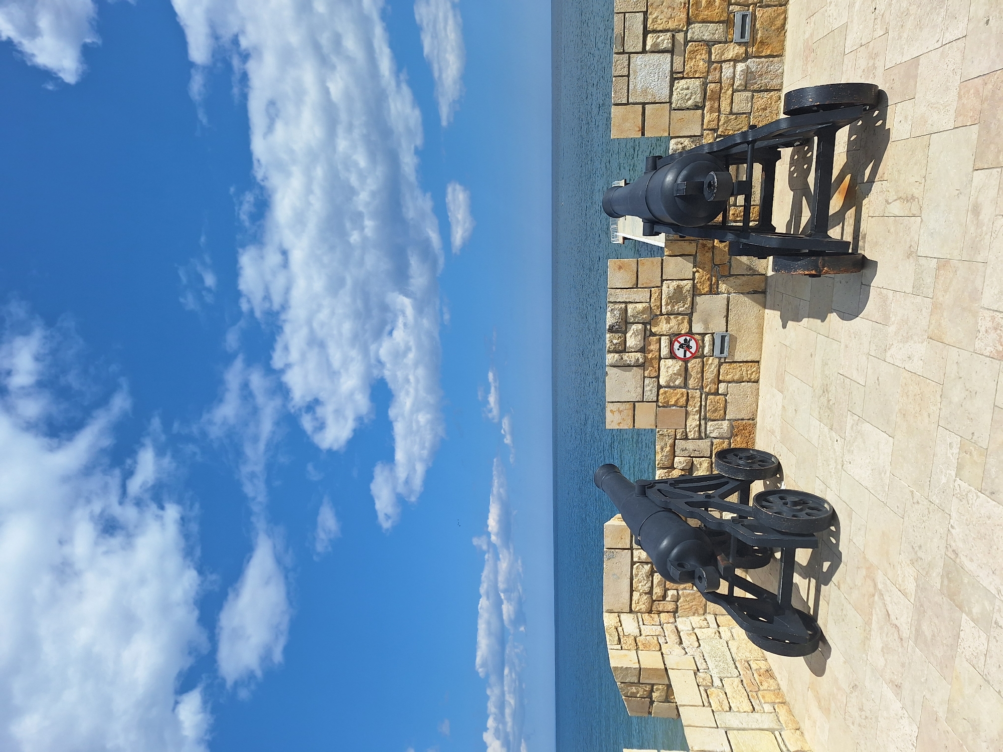 Two black cannons are mounted on a stone wall overlooking the sea in Varna, Bulgaria. The cannons are situated on a beige-colored tile surface near a stone wall constructed of light brown blocks. A sign with a red prohibition symbol is visible on the stone wall. In the background, the sea extends to the horizon, where it meets a blue sky dotted with white clouds.