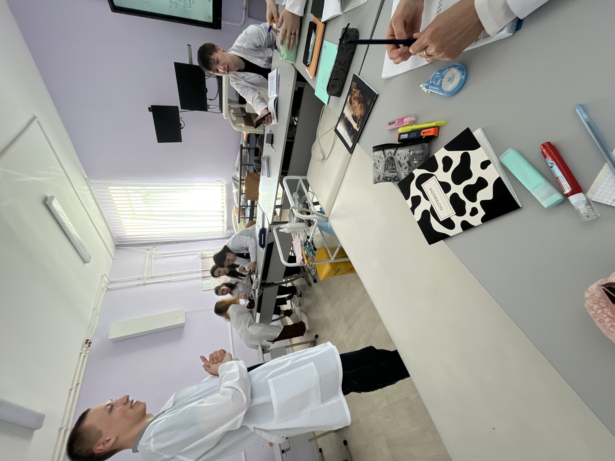 The image depicts a science classroom located in the city of Chaikovsky, Russia. The room has light purple walls, a white ceiling, and light-colored tiled floors. Fluorescent lights illuminate the room from the ceiling. A window with drawn blinds is visible along one wall.
Several students dressed in white lab coats are seated at long tables. Some are actively engaged in what appears to be a laboratory activity, interacting with notebooks and various stationery items such as pens, highlighters, and tape. A notebook with a black and white cow print design is visible on one table. A monitor is mounted on the wall near the ceiling at the end of the room where students are gathered. One male student is standing facing away from the camera with his arms crossed, seemingly observing the others.