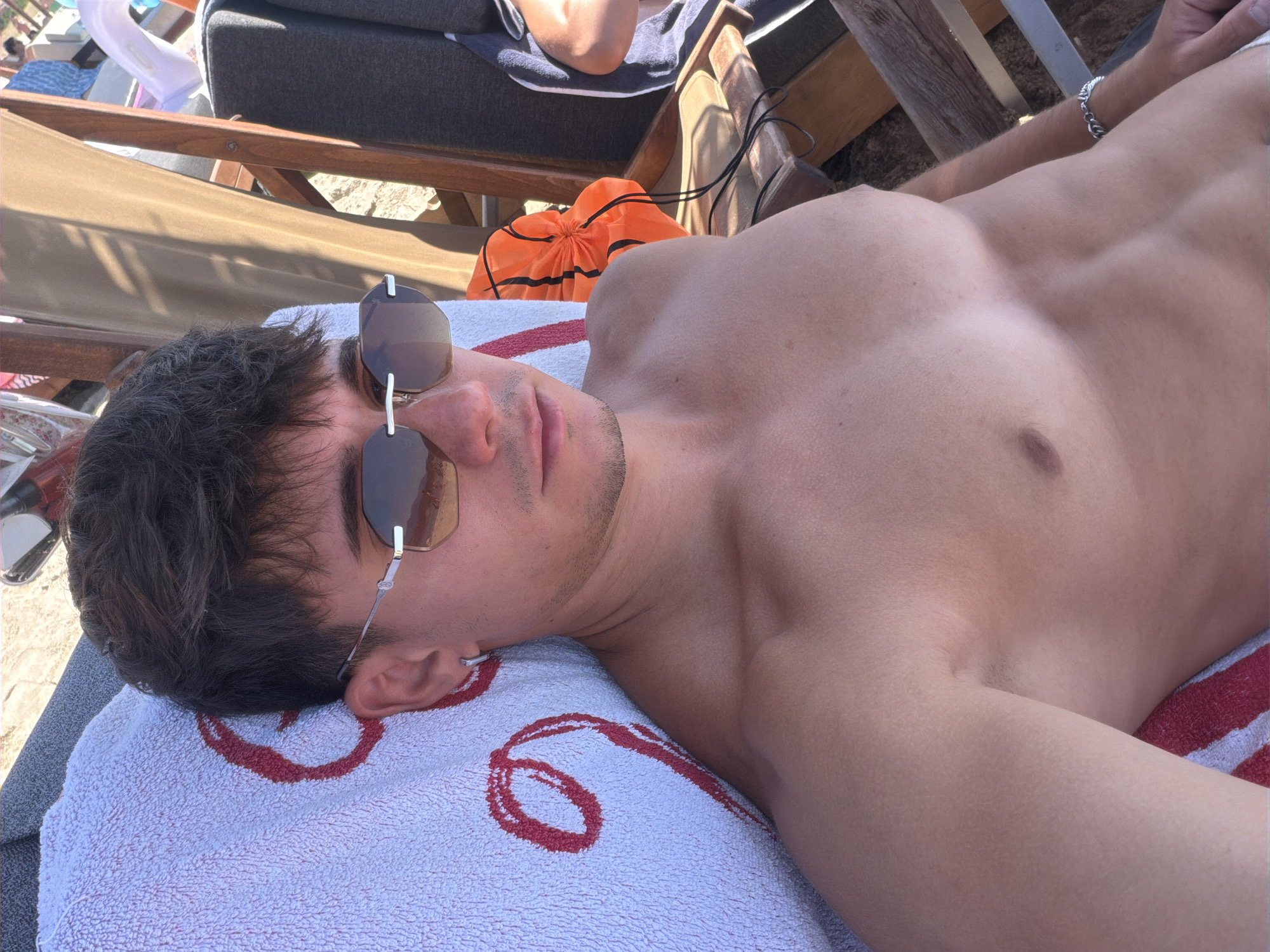 A young man with dark hair and sunglasses lies on a beach towel on a lounge chair at the beach. He is shirtless, wearing sunglasses, and has a silver earring in his left ear. The beach is sandy, and there are other lounge chairs and beachgoers visible in the background. The man is looking at the camera.