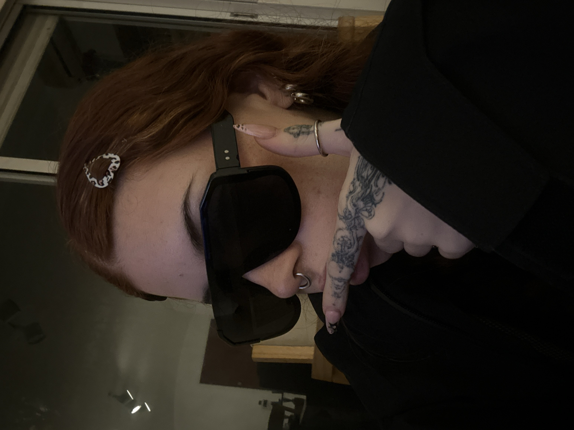 A close-up, low-angle shot shows a person with auburn hair, a fair complexion, and a nose ring. The person is wearing large black sunglasses and a black garment. A silver heart-shaped clip is visible in their hair. The person's left hand, adorned with a tattoo, is positioned near their nose. A silver ring is on their finger. The background includes a window and indistinct objects, suggesting an indoor setting. There is no information to suggest this photo was taken in Buenos Aires, Argentina.