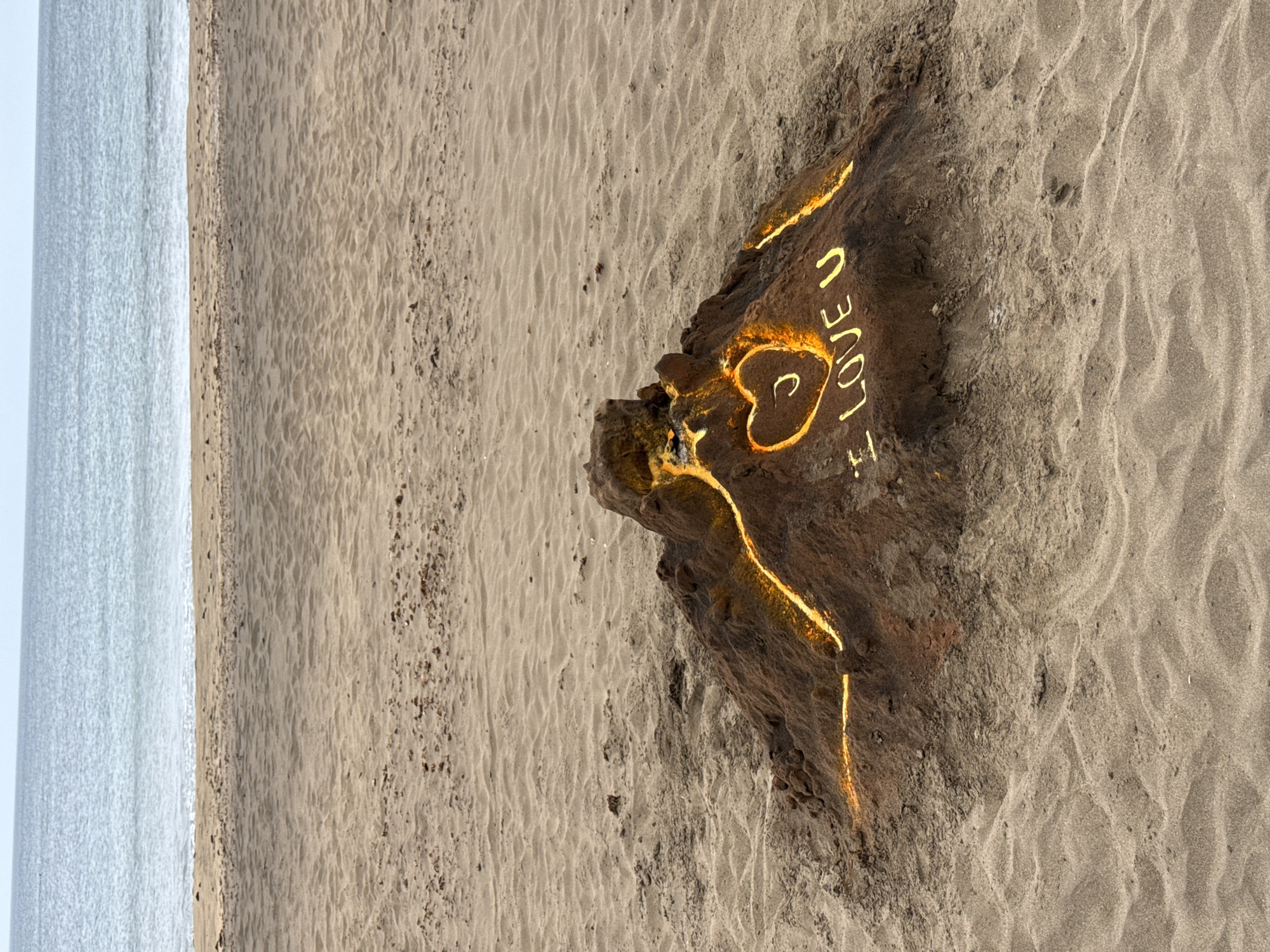 The image shows a beach scene, possibly near Tías, Spain. A large piece of driftwood lies on the sand, upon which the words "I LOVE U" have been written, with the heart symbol replacing the word "LOVE". The writing and parts of the wood appear to be illuminated or glowing with an orange or yellow light, suggesting either an artistic effect or the use of a light source. The beach stretches out to the side, and in the distance, what appears to be the ocean meets the horizon.