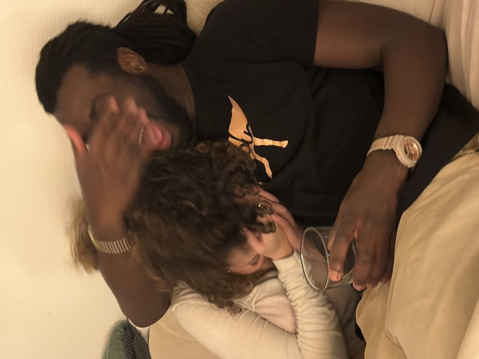 A man with dreadlocks, wearing a black t-shirt and a watch, is lying on a sofa with a woman. He has one hand over his face and his tongue is sticking out. The woman, with curly brown hair, is lying under him. She is wearing a light-colored shirt. A glass is held near her face. The man's other arm is resting on his leg, which is covered in khaki-colored pants.