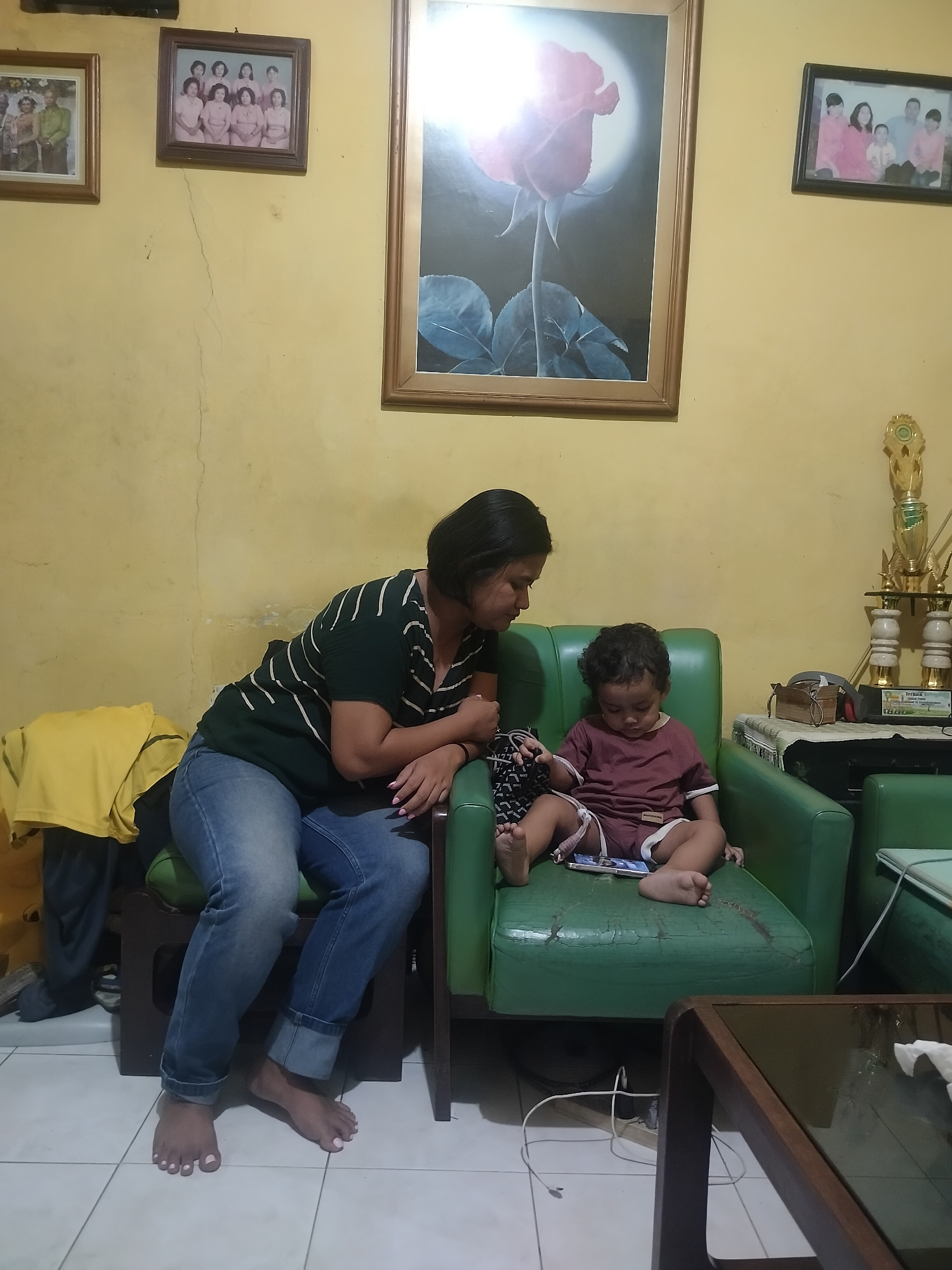 A woman and a child are sitting in a living room in Surabaya, Indonesia. The woman is wearing a dark green and white striped shirt and blue jeans. She is sitting on the left side of a green sofa, leaning forward. The child is wearing a maroon t-shirt and shorts and is sitting on the right side of the sofa, looking down at a phone. The walls of the room are painted yellow and have cracks. There are several framed pictures on the wall, including a family photo and a picture of a rose. There are also trophies on a shelf in the background. A glass-topped coffee table is in the foreground.