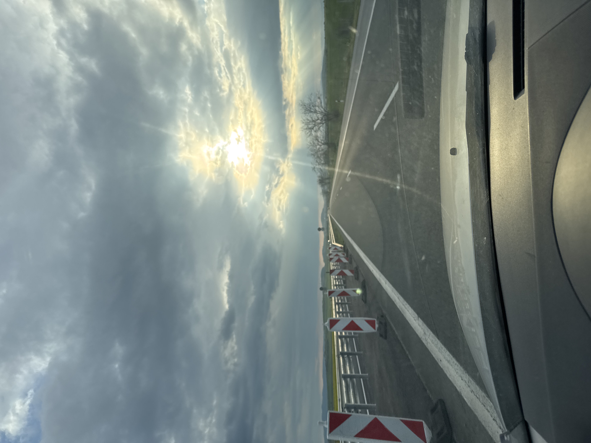 The image shows a view from inside a vehicle, possibly a car, traveling on a highway. The road has a white line marking the lane. Alongside the road, there are construction barriers with red and white striped markings. The sky is overcast with clouds and a bright spot indicating the sun. The landscape in the distance appears to be flat with some trees visible. There is no indication in the image that reveals that this scene is in the city of Chokoba, Bulgaria.