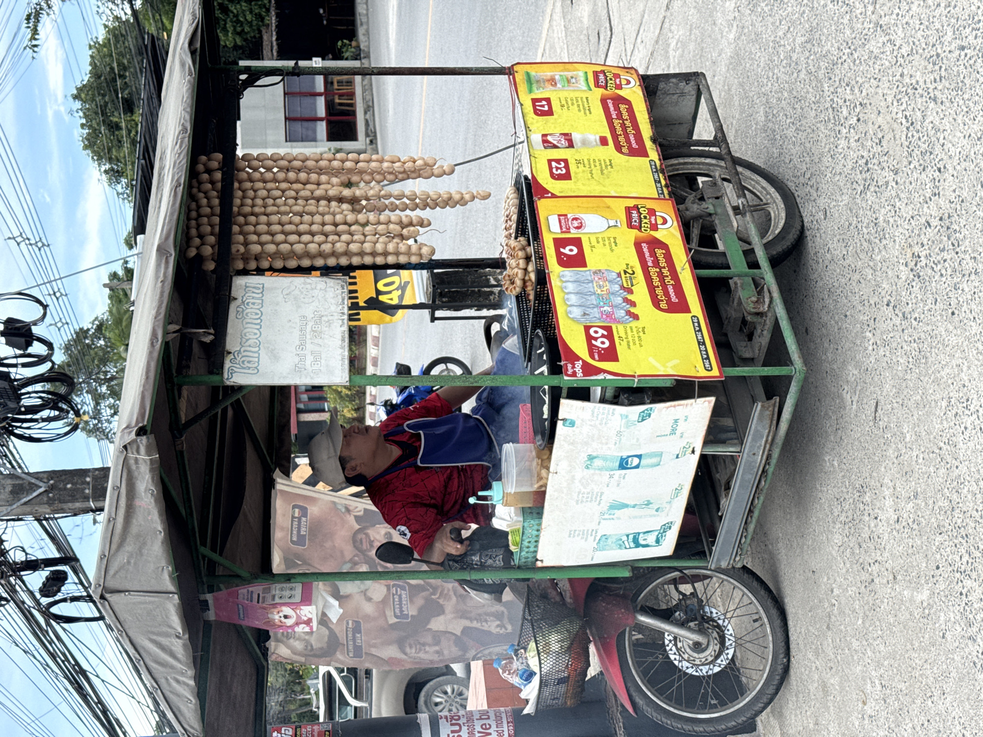 The image captures a street scene in Baan Bo Phut, Thailand, featuring a food cart operated by a man. The cart, which appears to be a modified motorcycle with a sidecar, is laden with goods. Prominently displayed are strings of sausages hanging from the canopy. The vendor, a man wearing a red shirt and a blue apron, is seated behind the cart, attending to a grill where sausages are cooking. Various bottles and containers are visible on the cart, suggesting a range of food and drink items. Colorful advertisements are affixed to the side of the cart, showcasing prices and promotions. The background shows a street with electrical wires overhead, suggesting an urban or semi-urban setting. Trees and buildings are also visible in the distance. The overall impression is that of a typical roadside food vendor operation in Thailand.