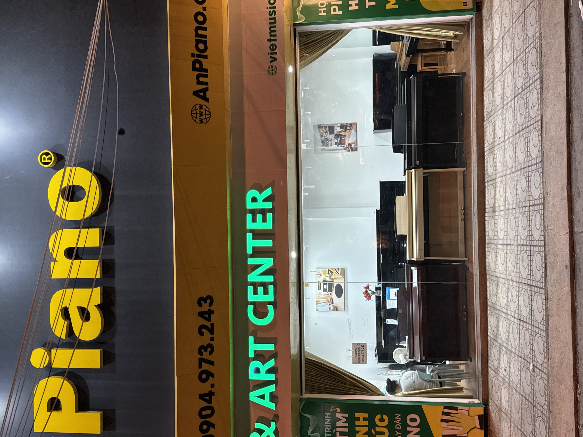 The image shows the storefront of "Piano & Art Center" located in Nha Trang, Vietnam. The exterior features a large, bright yellow "Piano" sign with a registered trademark symbol. Below this, a yellow banner displays a phone number "0904.973.243" and a website "AnPiano.com" with a globe icon. A green neon sign reads "& ART CENTER" with "vietmusic" and another globe icon to its right. The storefront has large glass windows displaying several digital pianos inside. Two women are visible inside the store, one sitting at a piano and another standing. The street in front of the store is paved with patterned tiles. A portion of a green sign with yellow text is visible on the left side of the image.