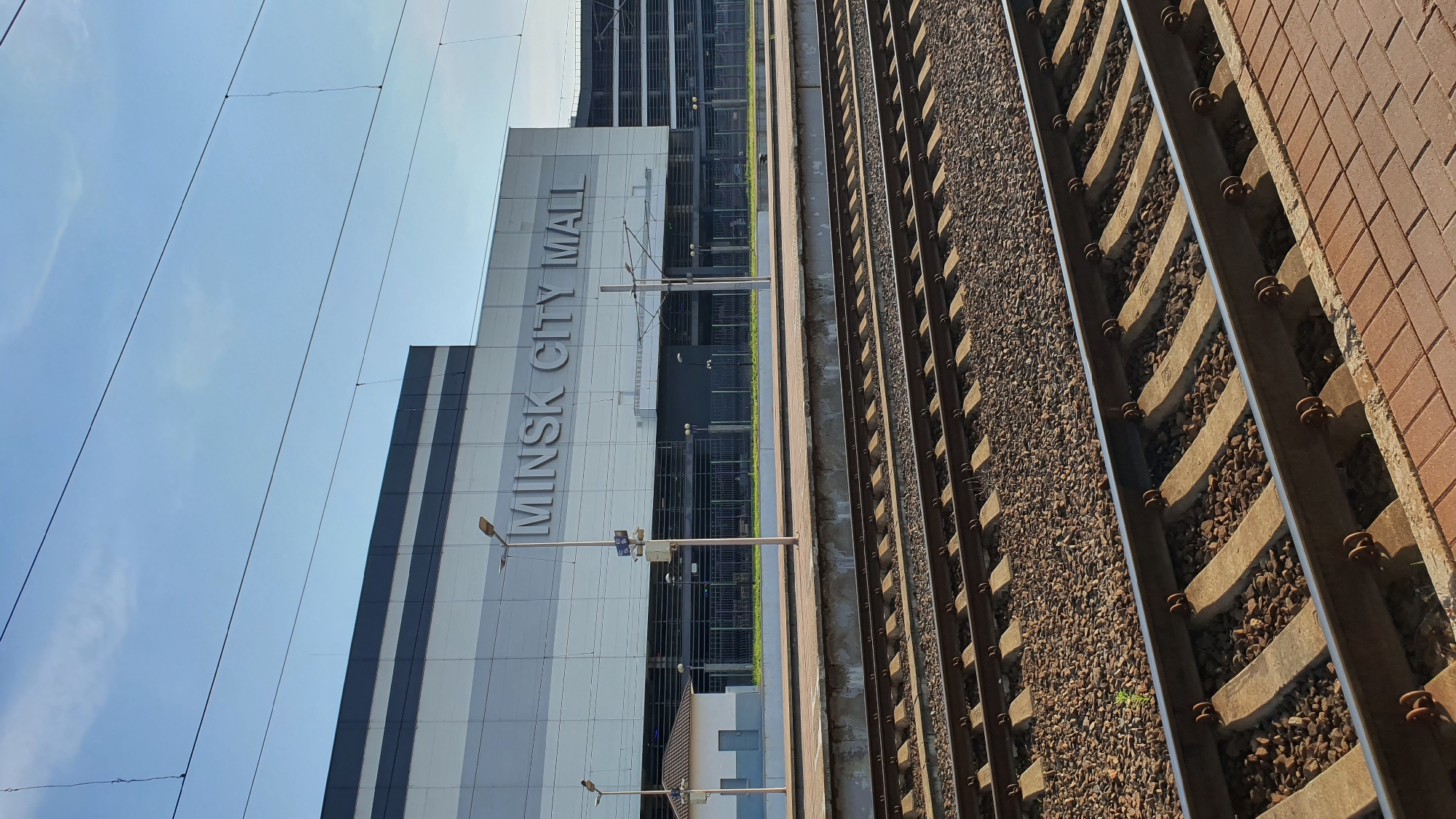 The image captures a view of the Minsk City Mall from a train platform. The mall's facade is prominently featured, with large, silver lettering spelling out "MINSK CITY MALL". The building appears modern, with clean lines and a light-colored exterior, accented by darker horizontal stripes. Several lampposts are visible along the platform in front of the mall.
To the right of the mall and platform, a set of train tracks is visible, running parallel to the platform. The tracks are made of metal rails laid on concrete sleepers, with gravel ballast beneath. The perspective is from a slightly elevated viewpoint, looking along the tracks and towards the mall.
The sky is a clear, bright blue with a few wispy clouds, suggesting it is daytime, likely late morning or early afternoon, with fair weather. The scene is that of a public space, a transportation hub adjacent to a commercial center. No people are visible in the frame, creating a sense of stillness. The overall impression is one of urban infrastructure and commercial development.
