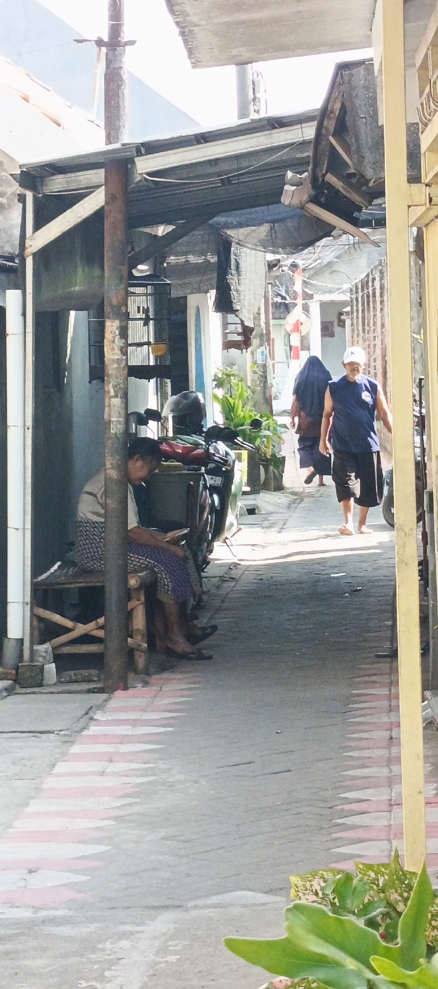 The image depicts a narrow alleyway in Krian, Indonesia, with overcast skies suggesting it might be a humid day. The alley is flanked by buildings on both sides, with makeshift roofing structures creating a shaded, somewhat cluttered environment. On the left, a person is seated on a wooden bench under a covered area, looking at a mobile phone. A motorcycle is parked nearby. Further down the alley, two figures are walking away from the camera. One is wearing a dark head covering and a long garment, while the other, a person wearing a blue t-shirt and shorts, has a white cap on. Green plants are visible in pots along the sides of the alley. The ground is paved with what appears to be bricks, with a pattern of red and white markings indicating a pedestrian crossing. The overall impression is of a densely populated urban area with elements of everyday life unfolding in a functional, albeit slightly rundown, setting.