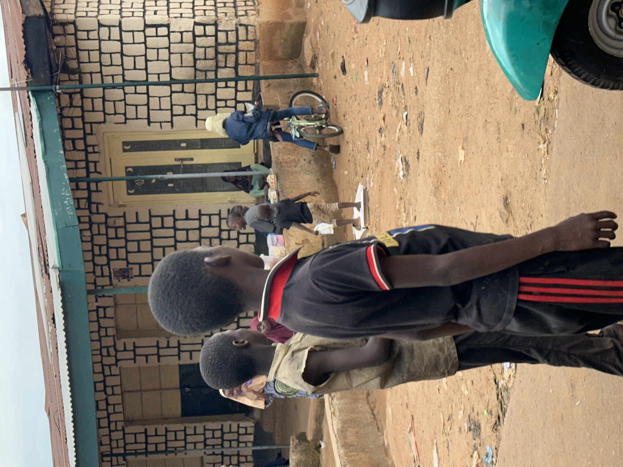 In Jos, Nigeria, the scene unfolds on a dusty street in front of a building with a corrugated metal roof and a facade of light-colored bricks with dark mortar. A young boy in a black t-shirt with red and white trim and black pants with red stripes stands in the foreground, his back to the camera. To his left, another boy in a light-colored, sleeveless shirt and dark pants is also facing away. In the middle ground, a child in a blue jacket and jeans is seated on a green bicycle. Several other children are visible near the building entrance. The ground is covered in reddish-brown dirt, scattered with debris. A turquoise scooter is partially visible on the right side of the frame. The sky is overcast.