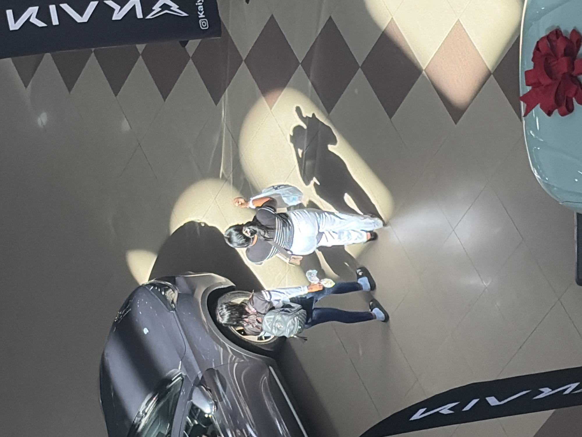 An overhead shot reveals an indoor scene, likely a car showroom or exhibition, in Ciudad Guayana, Venezuela. The floor is covered with light-colored tiles featuring a repeating pattern of dark and light diamond shapes. Bright, directional lighting from above casts long, distinct shadows of the subjects onto the floor.
Two individuals are visible, observed from a bird's-eye perspective. The person positioned further away from the camera wears a striped top, light-colored pants, and has dark hair pulled back, with a light-colored bag slung over their shoulder. The other person, closer to the camera, is dressed in a dark top, dark pants, and a backpack, holding a clear plastic bottle. They appear to be casually observing the displayed vehicles.
Two cars are partially visible. On the left, a dark grey or black car's front quarter is seen. To the right, a light blue or turquoise car stands out, adorned with a large, vibrant red bow on its front, indicating a special presentation or recent purchase.
Black banners with white text are prominent in the scene. One banner at the top clearly displays "KIVVA" alongside a stylized logo resembling a mountain range or tree, with "@Kaly" also visible. Another banner bearing the "KIVVA" text is seen at the bottom right of the frame. The overall atmosphere suggests an active display area within a commercial space.