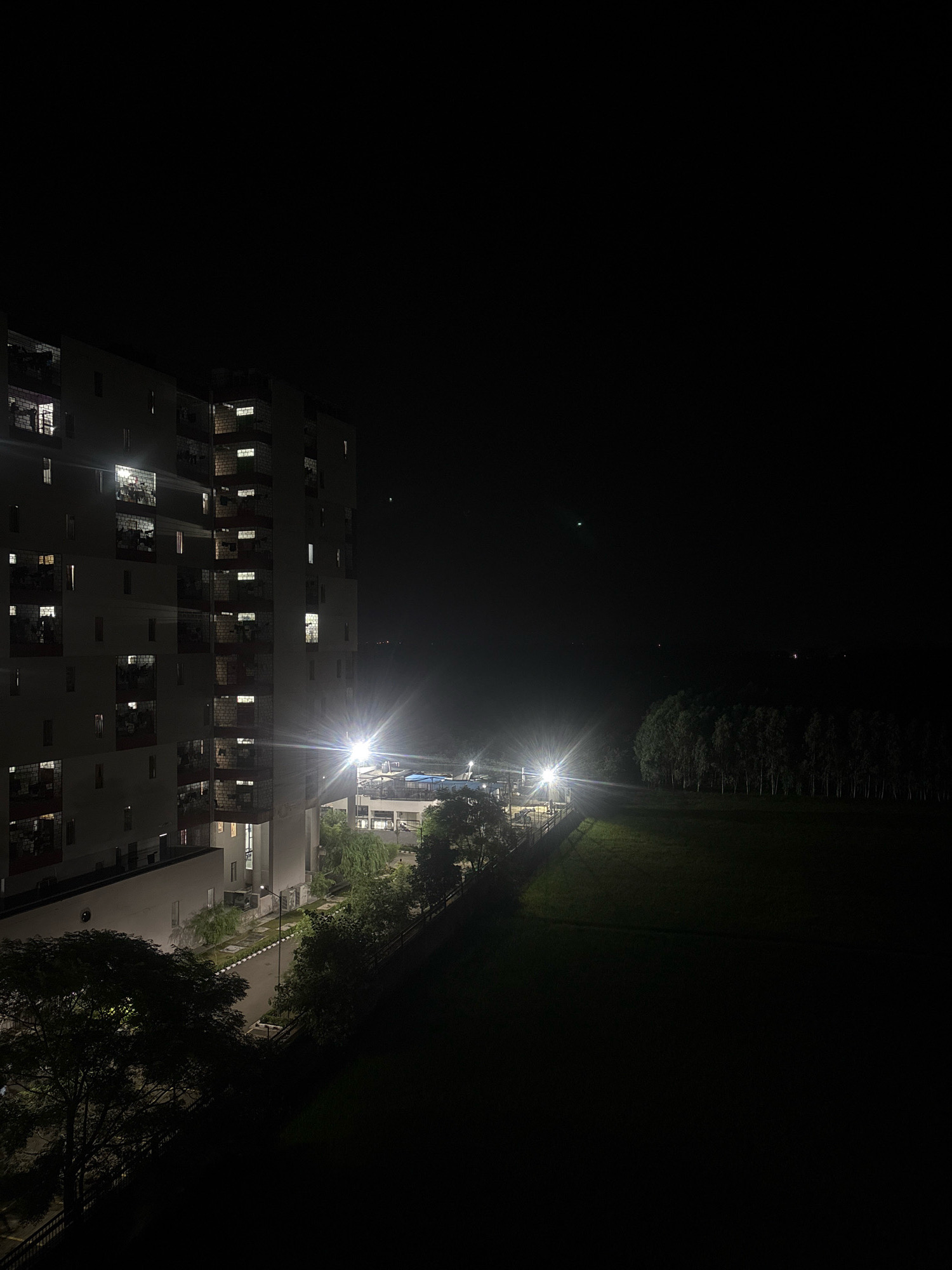 This is a nighttime, wide-angle, outdoor shot of a residential apartment building in Gharuan, India. The building is tall and modern, with multiple floors featuring balconies and numerous windows, many of which are illuminated, suggesting occupancy. The building is located adjacent to a dark, grassy field. Several bright, artificial lights illuminate the area around the building and the field. A line of trees is visible in the distance, bordering the field. The sky is dark and clear, with a few faint, distant lights. The overall atmosphere is calm and serene, with a hint of urban life contrasted against a natural backdrop.
