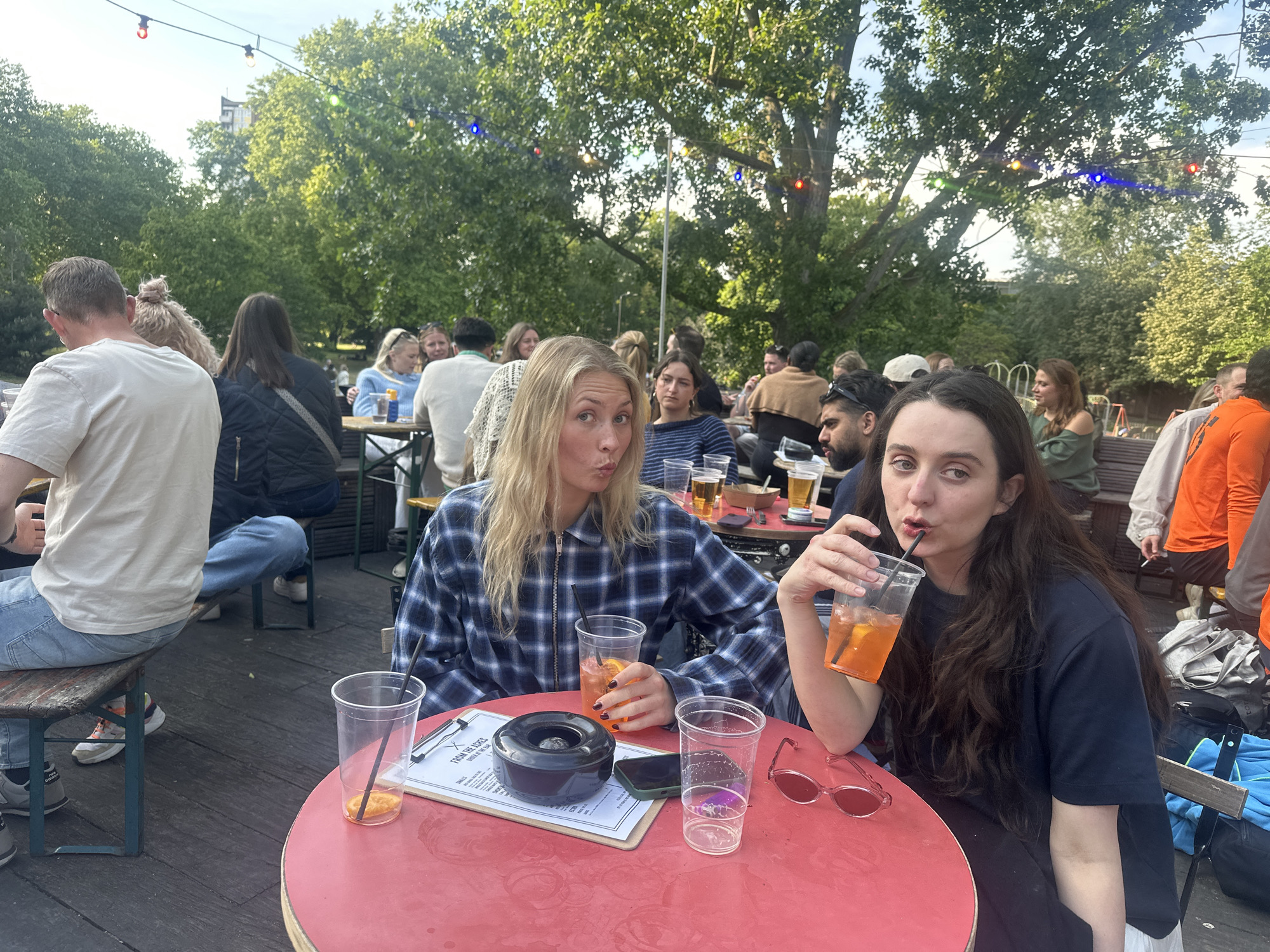 The image depicts an outdoor scene in what appears to be a beer garden or patio, likely in London, United Kingdom. Several people are seated at picnic-style tables. Two women are seated at a small, round red table in the foreground. The woman on the left has long blonde hair and is wearing a blue plaid zip-up shirt. She is holding a plastic cup with an orange-colored drink and is making a pouty face. The woman on the right has long dark hair and is wearing a dark blue shirt. She is drinking from a similar plastic cup with a straw and making a similar expression. On the table, there is an ashtray, a menu on a clipboard, a cellphone, sunglasses, and additional plastic cups. The background includes other patrons sitting and standing, trees, and string lights overhead. The setting suggests a relaxed, social atmosphere.