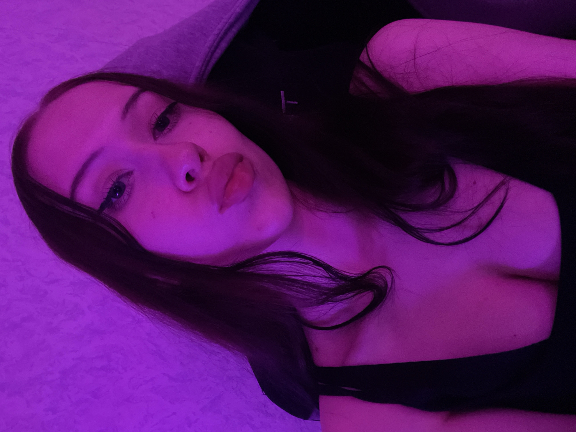 A young woman with dark hair and fair skin is pictured in a close-up shot, with her face angled towards the camera. She is wearing a black top, and her hair is long and dark, falling around her shoulders. The background is a textured purple wall. The lighting is predominantly purple, casting a distinct hue over the scene. The woman's lips are slightly parted, and she appears to be looking directly at the camera. The image appears to be taken indoors, possibly in a bedroom or personal space. No specific details about Votkinsk, Russia, are visible in the image.