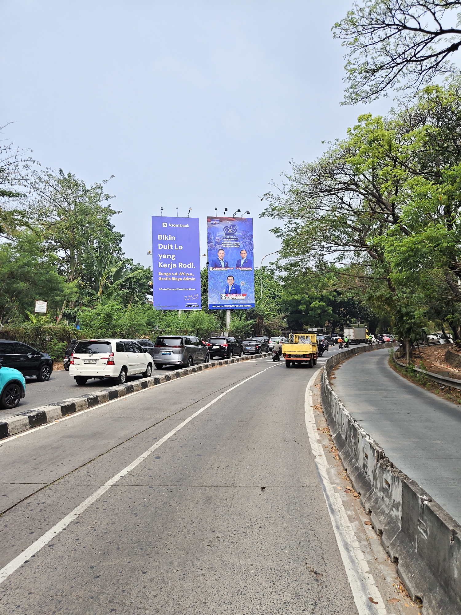 The image depicts a traffic scene on a multi-lane road in South Jakarta, Indonesia, likely along Jl. TB Simatupang, based on the presence of billboards for "Krom Bank" and the general urban landscape. The road is paved with asphalt and features white lane markings. A concrete barrier separates the lanes from the right side of the road, which is bordered by dense green trees and vegetation. Billboards are prominently displayed on the left side of the road, advertising "Krom Bank" with slogans in Indonesian. The billboards feature images of two men in suits, one of whom appears to be a formal portrait, and the other engaged in a handshake.
Traffic is moderate, with several vehicles visible, including white SUVs, a light blue car, black sedans, a yellow flatbed truck, and a white truck. Motorcycles are also present, weaving through the traffic. A few individuals can be seen on motorcycles. The sky is overcast, suggesting a bright but cloudy day. The overall environment is a typical urban setting characterized by road infrastructure, vehicular traffic, advertising billboards, and abundant greenery.