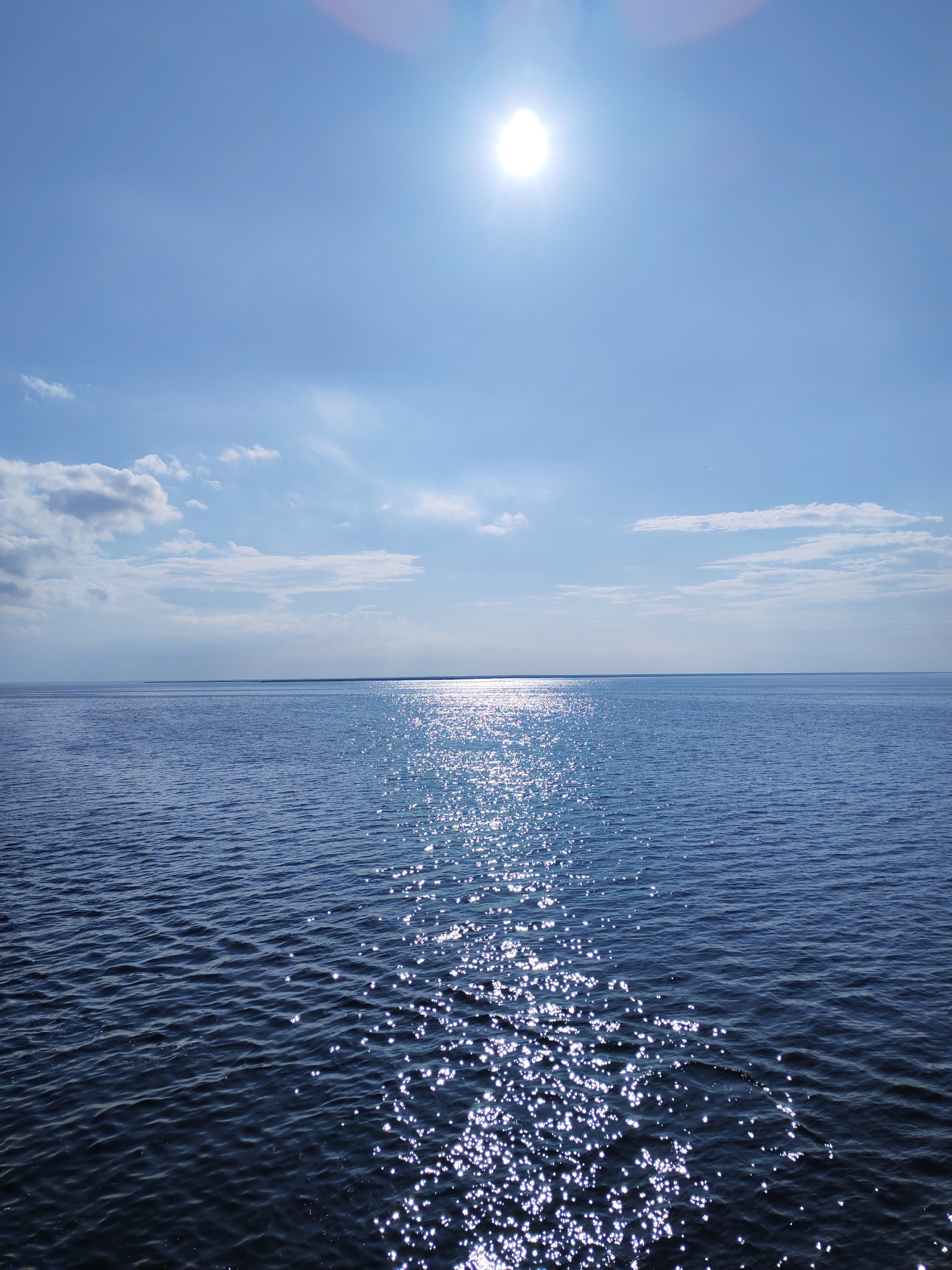 The image depicts a body of water, possibly the Volga River, reflecting the sunlight. The sky is blue with some scattered white clouds. The sun is high in the sky, creating a bright glare. The water surface is rippled, and the reflections of the sun appear as scattered bright spots. The horizon line is visible in the distance. Based on the context of Mologa, Russia, a city submerged in 1941 after the creation of the Rybinsk Reservoir, this could be a view of the Rybinsk Reservoir with only the horizon line visible. There are no people or other recognizable objects in the image.