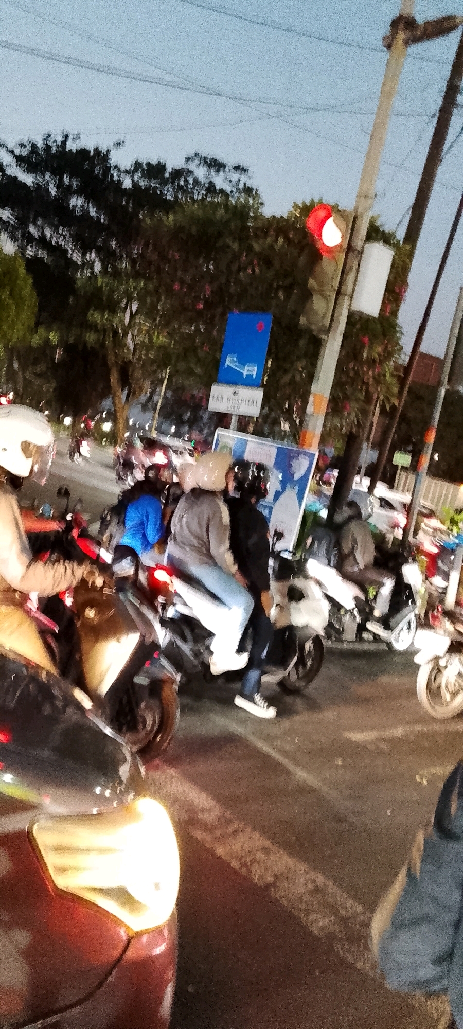 In Depok, Indonesia, at dusk, a traffic scene unfolds at an intersection. Multiple motorcycles are present, some carrying passengers. In the foreground, the headlight of a red car is illuminated. A traffic light, with its red signal illuminated, is visible above the intersection. Road signs are also present, including one indicating "EKA HOSPITAL 1.5KM" and another blue sign with an icon of a bed. The sky is a dusky blue, and trees line the street. The overall impression is one of urban traffic during the evening transition.