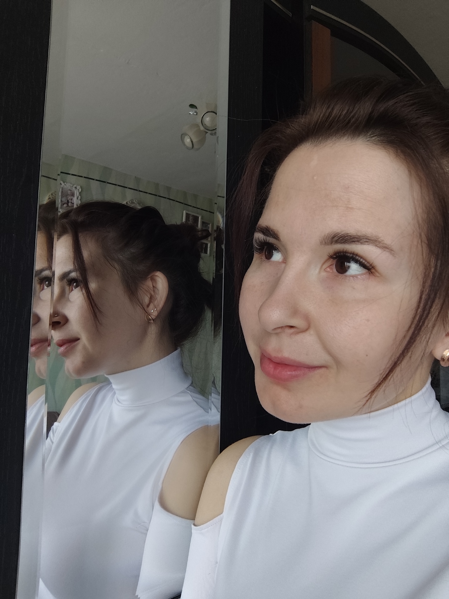 An indoor portrait features a woman posing, captured directly in the foreground and as a reflection in a mirror to her left. The woman, with dark brown hair styled partially pulled back and loose strands framing her face, is seen from the chest up. She is looking slightly upward and to her right with a gentle, closed-mouth smile, her expression appearing serene and contemplative. She wears a white, high-neck top with cut-out shoulders and a small gold-colored stud earring in her right ear. Her eyelashes appear long, possibly enhanced.
To the left, a large mirror with a dark frame reflects her profile, showcasing a similar pose. The reflection also reveals parts of the room behind her, including a light-colored ceiling with two distinct light fixtures. Below the ceiling in the reflection, a wall is visible, decorated with greenish-striped wallpaper and featuring a white-framed picture. A dark door frame is partially visible in the upper right of the reflected scene. The setting suggests a home environment, likely in Фоки, Russia. The lighting is soft and even, highlighting her features. There is no visible text in the image.