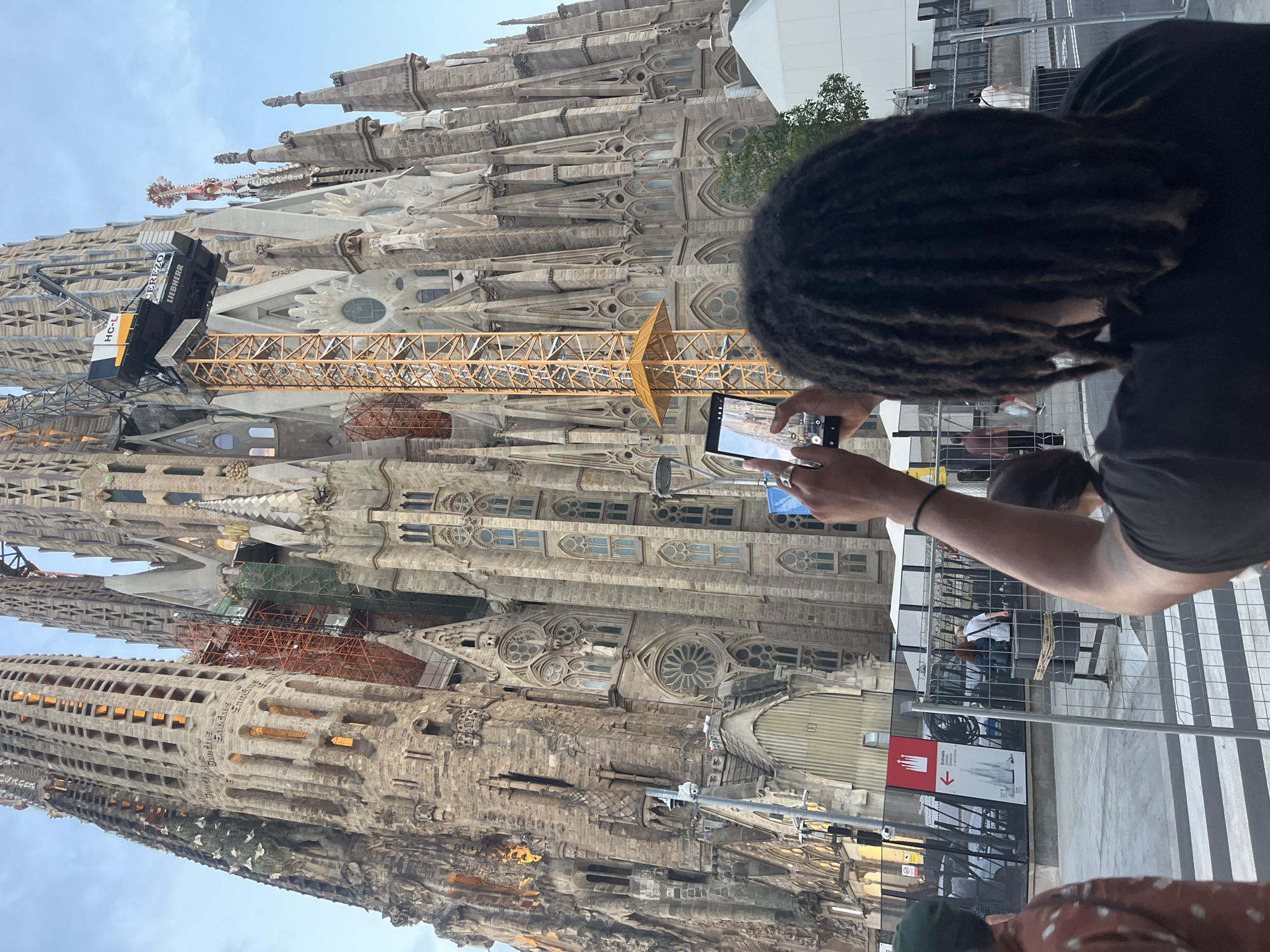 The image shows a person with dreadlocks in the foreground, holding a smartphone to take a picture of the Sagrada Familia basilica in Barcelona, Spain. The basilica is a large, ornate stone structure with intricate carvings and spires, and a crane is visible at the top, indicating ongoing construction or renovation. The sky is a pale blue with some clouds. In the background, a chain-link fence separates the viewer from the building, and other people can be seen milling around. There are also signs visible, one of which appears to be directional. The overall lighting suggests it is either early morning or late afternoon.