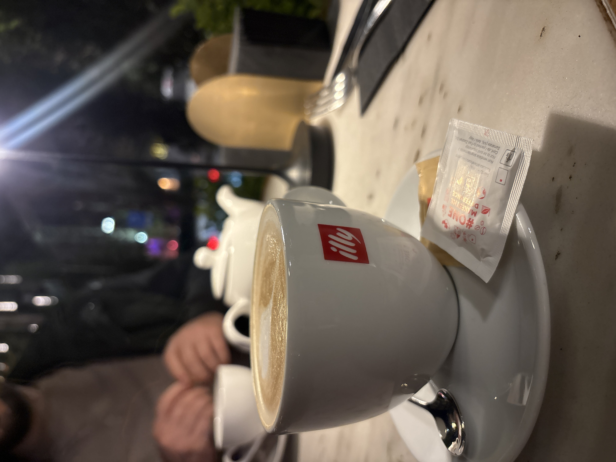 The image presents a warm, low-light scene at a cafe in Yerevan, Armenia, likely captured during the evening or night. The main subject is a white coffee cup, prominently featuring the red "illy" logo, filled with a beverage displaying delicate latte art. The cup rests on a matching white saucer with a spoon. A small, white sachet of sweetener is placed on the saucer next to what appears to be a small pastry or biscuit.
In the softly blurred background, a white teapot and another smaller white cup are visible, suggesting a shared coffee or tea experience. Partially visible hands belonging to another person can be seen in the mid-ground, adding to the impression of a social setting. The table surface is a light, possibly marble, material, reflecting some of the ambient light.
The distant background is dark and out of focus, featuring various bokeh lights, indicating streetlights or indoor lighting from other establishments, confirming the nighttime atmosphere. Some blurred, golden-toned objects are also present in the mid-ground, possibly decorative elements or light fixtures within the cafe.
Notable text includes the "illy" brand on the cup. The sweetener sachet displays text in Italian and English: "#ONE MAKES THE DIFFERENCE", "Raccolta differenziata / Non vendibile singolarmente / Not to be sold separately", and "Conf. da / packed by Fast Zuccheri S. Samarate (VA), Italia / Italy". The scene conveys a calm and relaxed atmosphere, ideal for enjoying coffee or tea.