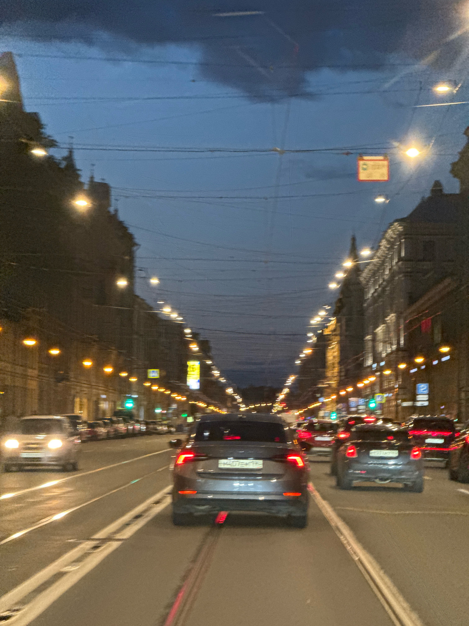 The image depicts a street scene in Saint Petersburg, Russia, at dusk or early night. The perspective is from inside a vehicle, looking forward. The street is lined with ornate buildings illuminated by streetlights and lights from within the buildings. Overhead, a complex network of electrical wires, likely for trolleybuses or trams, crisscrosses the sky. The sky is a deep blue with dark clouds visible.
Several cars are present on the road, appearing to be in traffic. The foreground features the back of a dark SUV, its taillights illuminated. To the left, a silver car with its headlights on is visible. Further down the street, more vehicles and the glow of traffic lights can be seen. The presence of tram tracks on the road is also noticeable. A rectangular sign with green and white markings is suspended overhead. The overall impression is of a busy urban environment at the transition from day to night.