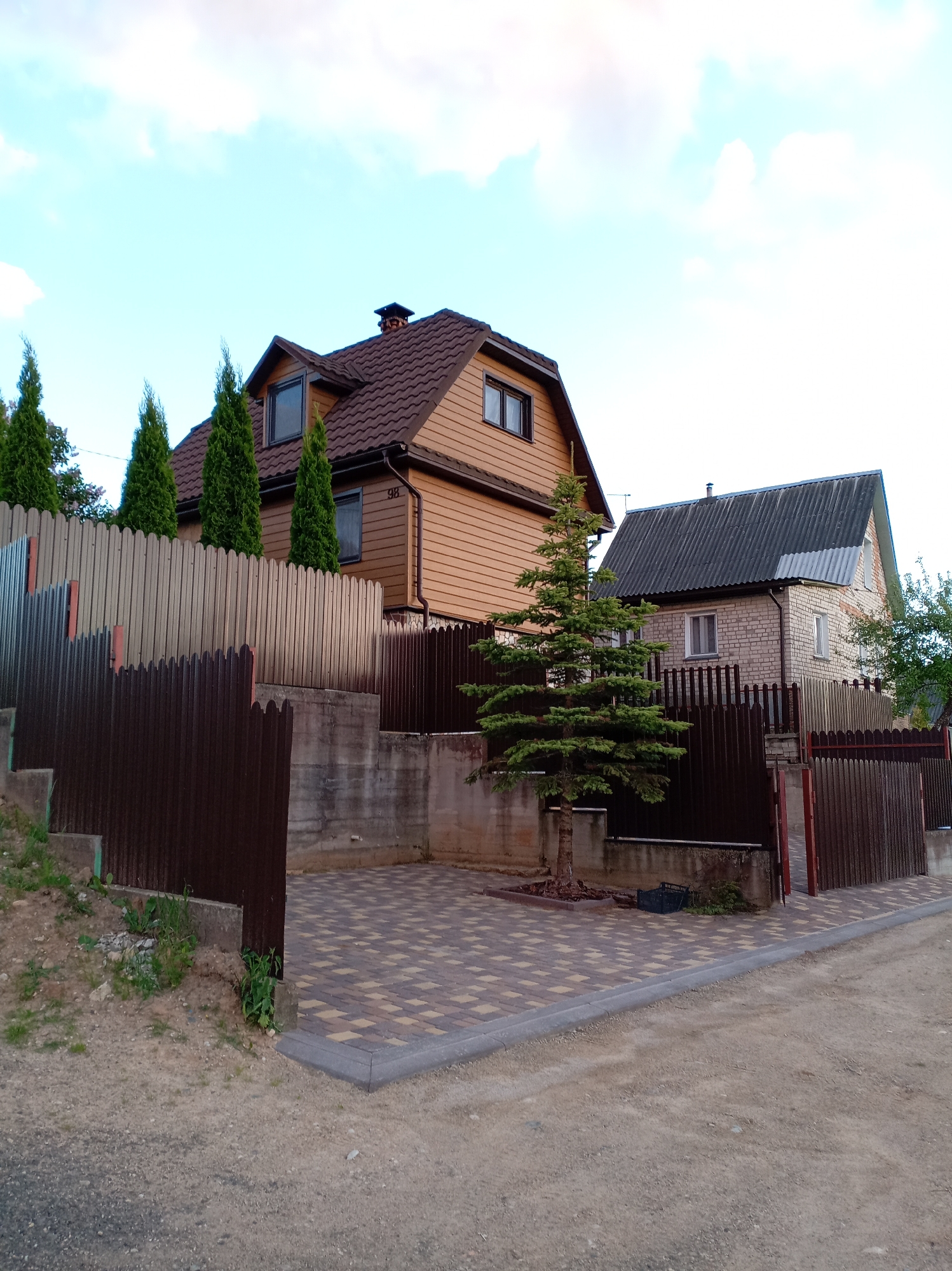 The image shows two houses behind a fence. The house on the left has brown siding and a brown shingle roof. The house has a chimney and two dormer windows. There are four evergreen trees in front of the fence. The house on the right has light-colored brick siding and a gray shingle roof. The fence is made of dark brown metal. The ground in front of the fence is covered in brown and yellow paving stones. There is a dirt road in the foreground. The sky is blue with some clouds. The location is a residential area. The city is unknown.