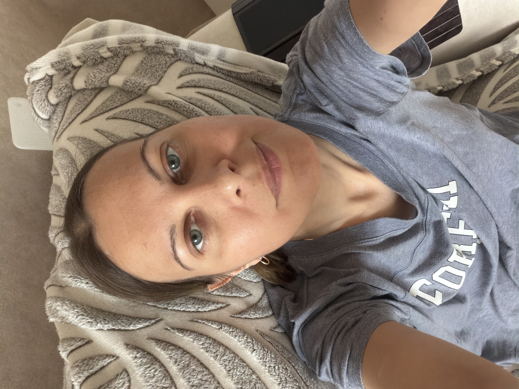 A woman is lying down on a couch, possibly at home. She is wearing a grey t-shirt with white lettering. The couch has a textured blanket draped over it. The scene appears to be indoors, in a residential setting. There is no indication of the specific location within Izhevsk, Russia.