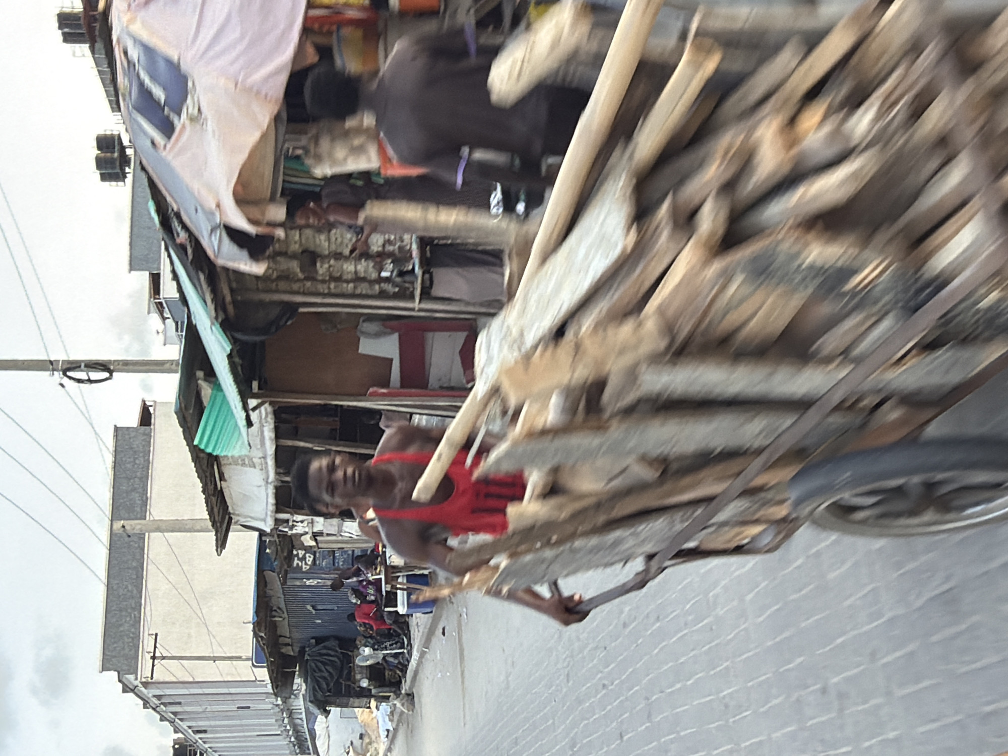 The image depicts a man in Igboefon, Nigeria, pulling a cart loaded with lumber along a paved street. The street is lined with small shops and dwellings, some with corrugated metal roofs. Other people are visible in the background, some seated. Overhead, power lines are strung between poles. The sky is overcast.