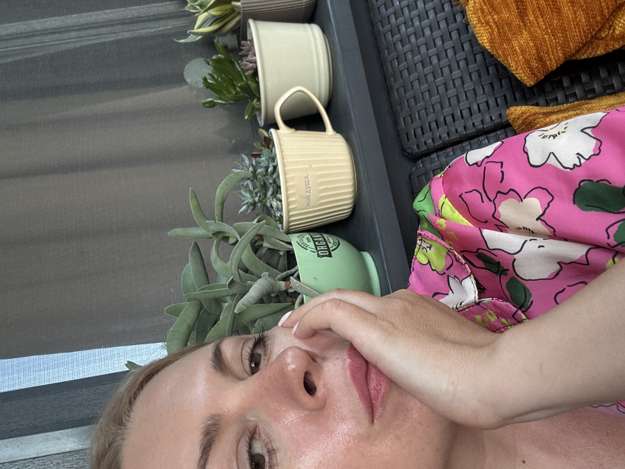 An eye-level, medium-shot photograph shows a fair-skinned woman with blond hair in a pink and white floral shirt. She is resting her right hand on her cheek, with her fingers extended towards her eye. She is looking to the left of the frame. Behind her, a row of potted succulents is visible on a dark shelf. The plants are of various shapes and sizes, with some in green, tan, and striped pots. A gray, woven blind or curtain is in the background. The overall impression is one of a relaxed, outdoor setting, possibly a balcony or patio. The presence of the succulents and the woven furniture suggests a warm climate, potentially consistent with Sochi, Russia.