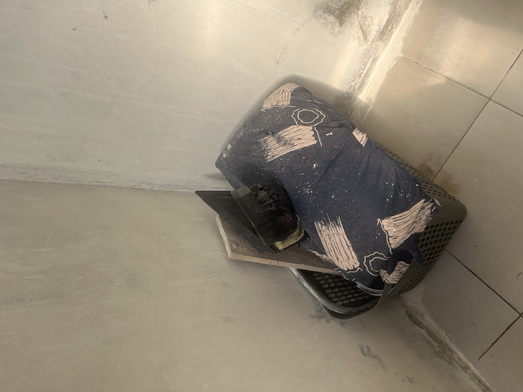 The image shows a dimly lit interior corner of a room, likely undergoing renovation. The walls and floor are tiled in a neutral tone and appear dusty and unfinished. In the corner, a dark grey plastic basket is filled with what appears to be construction debris, including a wooden plank and a black tool. A folded dark blue fabric with a light pink abstract pattern is draped over the contents of the basket. The lighting suggests it may be evening or an area with limited natural light. This scene does not contain any elements that would indicate a specific location in Cirahayu, Indonesia.