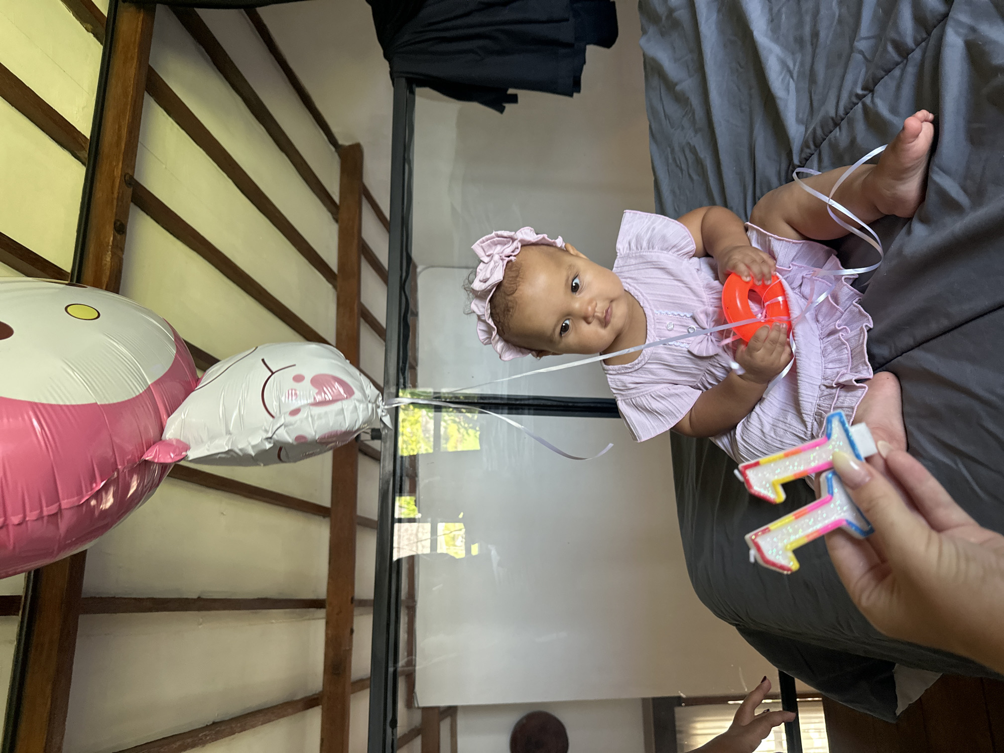 In North Kuta, Indonesia, a baby girl is seated on a bed. She is wearing a light purple dress and a pink headband. Two balloons, one pink and white with a Hello Kitty design and another white with paw prints, are visible above her. In the foreground, a hand holds two candles shaped like the number "11". The room has a sloped ceiling with wooden beams and appears to be indoors.