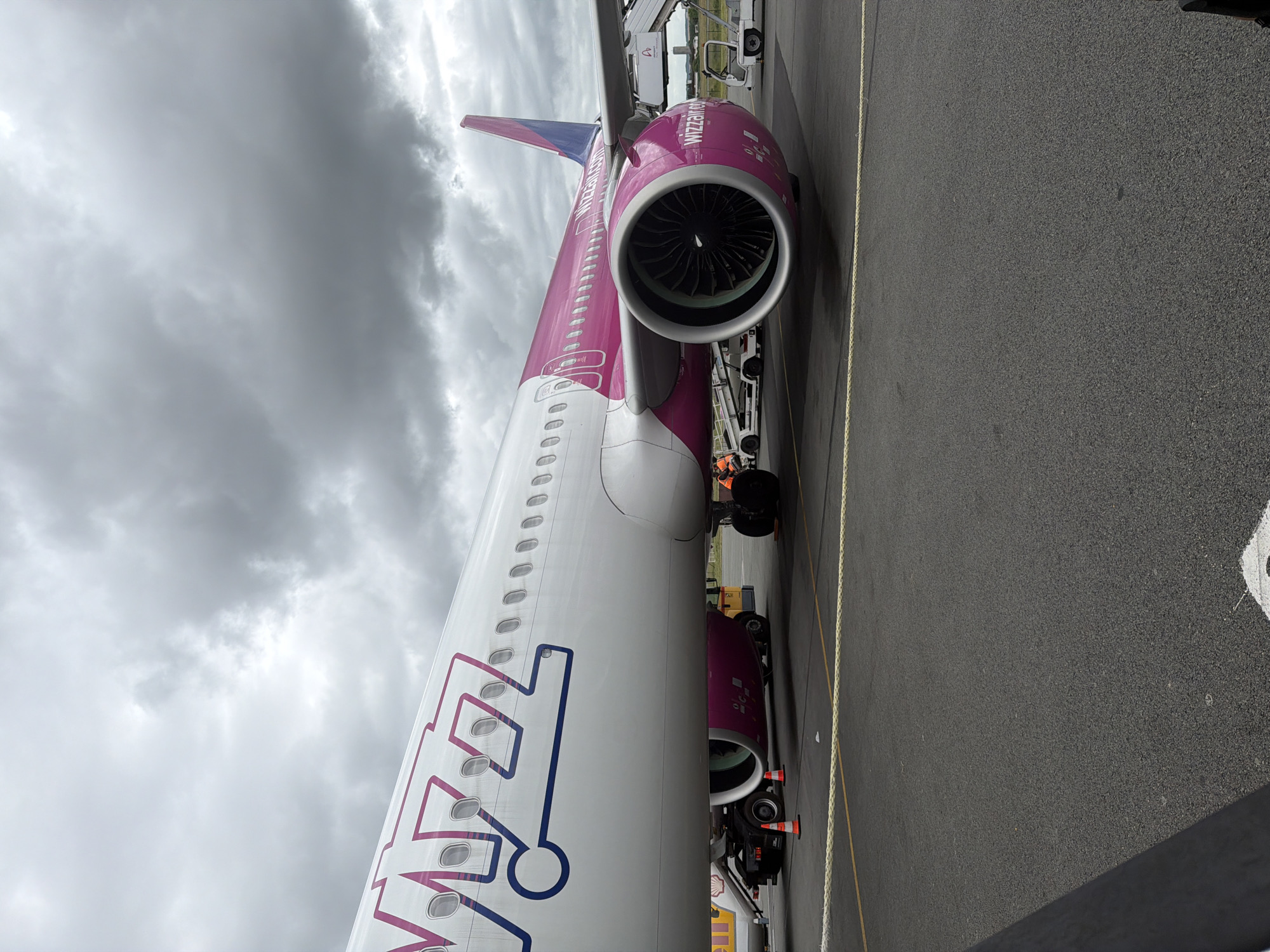 A Wizz Air Airbus aircraft is prominently featured, parked on an airport tarmac under a heavily overcast sky. The airplane displays its distinctive white fuselage with purple accents on the tail, engine nacelles, and a stylized "WIZZ" logo on the forward section of the fuselage.
The image is captured from a slightly elevated perspective, looking along the left side of the aircraft towards its rear. The large jet engine is visible beneath the wing, its fan blades clearly distinguishable within the engine's intake. The text "Wizz Air" and "wizzair.com" are inscribed in white on the purple engine cowling. Further back, the purple vertical stabilizer (tail fin) features "WIZZ AIR" written vertically. Numerous passenger windows line the aircraft's side.
On the ground, an airport jet bridge or mobile stairs is positioned near the aircraft's boarding door, and various ground support equipment, including what appears to be a fuel truck and other service vehicles, are visible near the plane's undercarriage and engine area. The tarmac is dark asphalt with yellow painted lines indicating boundaries or pathways. The sky is filled with thick, grey clouds, suggesting an overcast daytime.
The scene depicts typical ground operations around a parked commercial passenger jet at an airport, identified as being in Tillé, France, which corresponds to Beauvais-Tillé Airport.