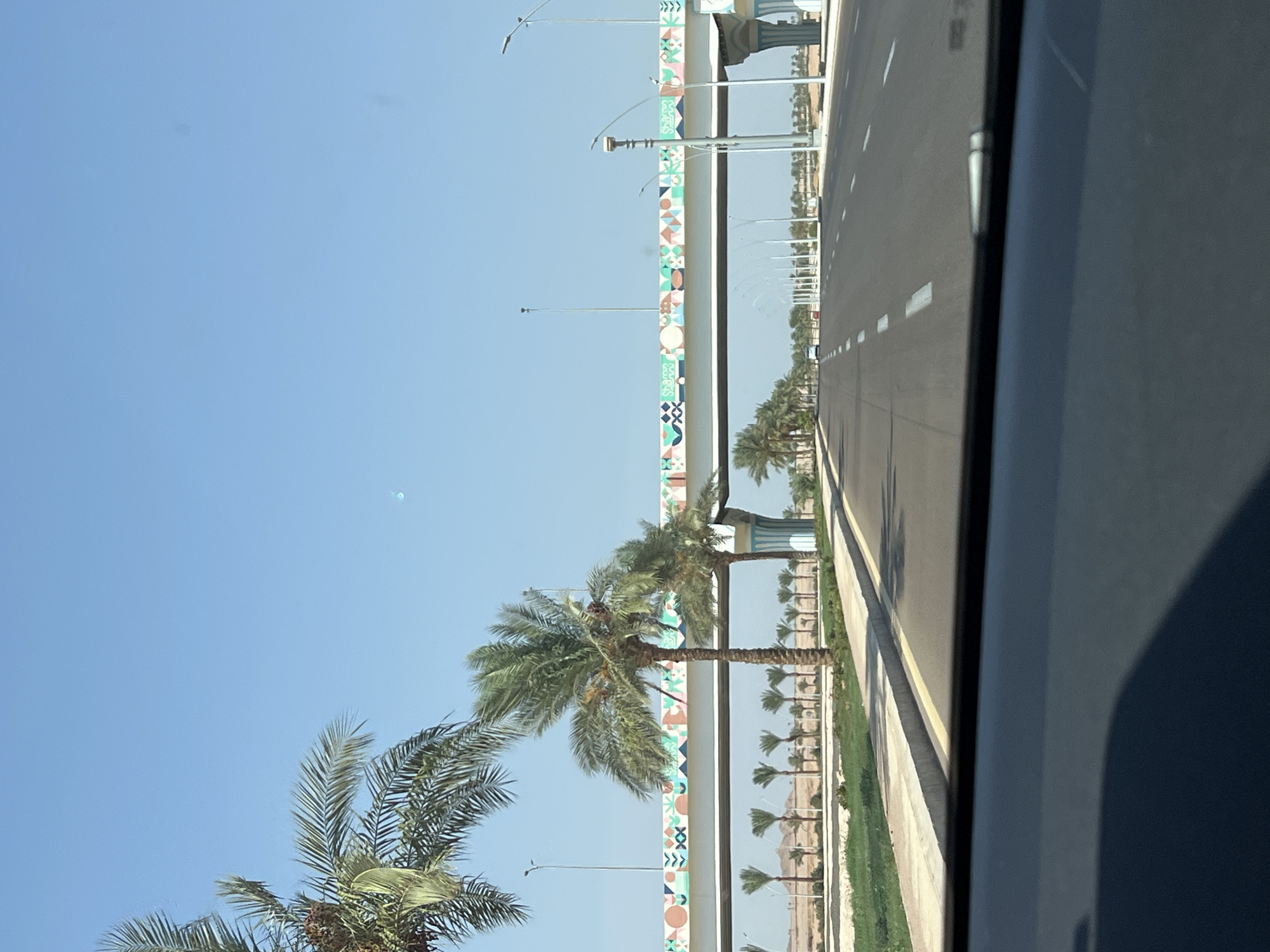 The image shows a wide, paved road leading into the distance under a clear blue sky. Along the sides of the road, palm trees are planted in neat rows, and a concrete barrier with a decorative, geometric pattern runs across the top of an elevated structure spanning the road. Streetlights are visible along the median and sides of the road. The ground on either side of the road is covered in sand and low-lying vegetation, with some distant buildings or structures visible in the background. The view appears to be from the interior of a vehicle, as the dashboard is visible in the foreground. The scene depicts a modern infrastructure development in a sunny, likely arid, environment.