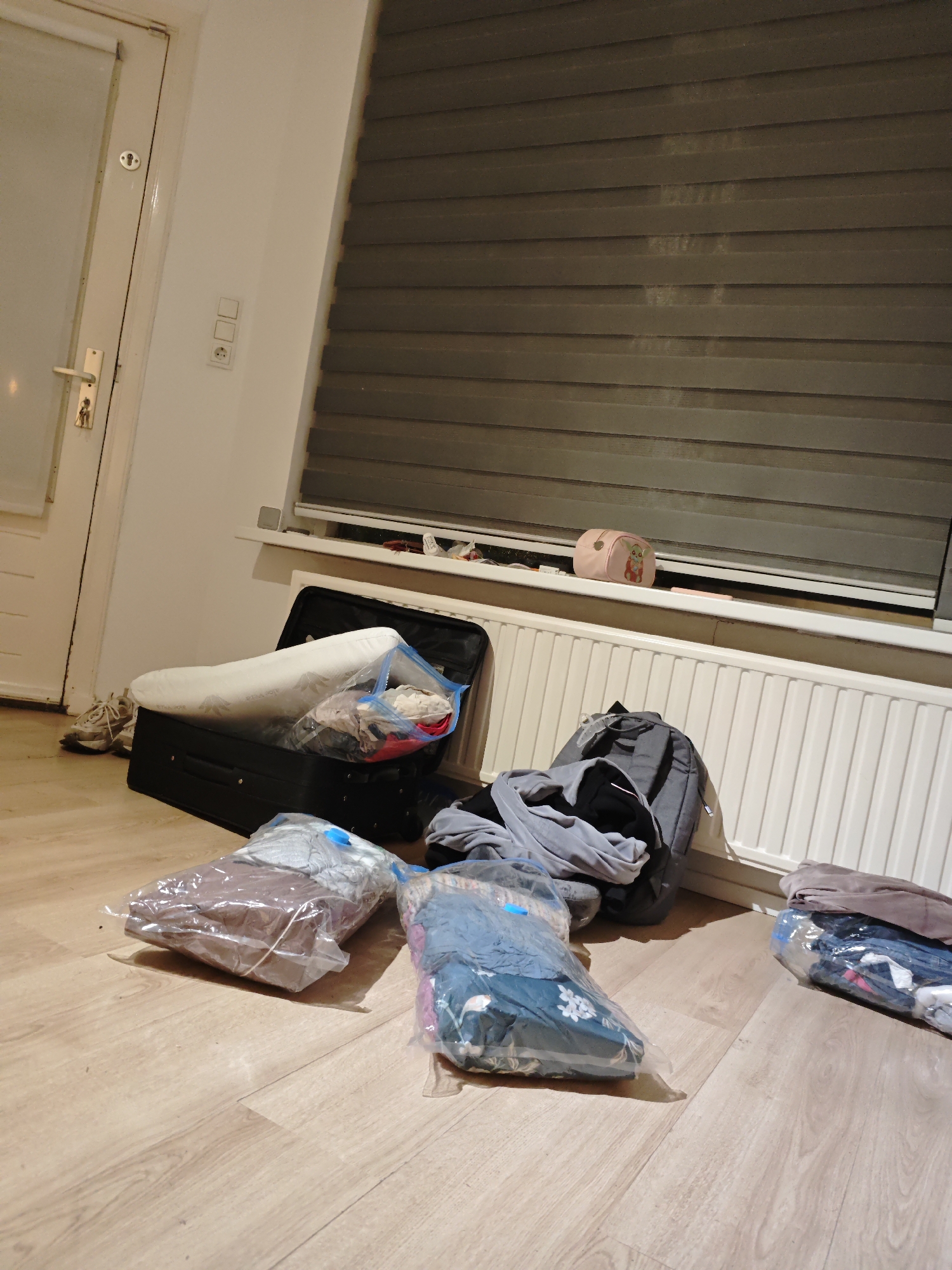 In a room, likely in Eindhoven, Netherlands, a black suitcase lies open on a light brown wooden floor. The suitcase contains a white pillow and several items packed in clear plastic bags. Adjacent to the suitcase, on the floor, is a grey backpack and various other plastic bags filled with clothing. To the left of the suitcase, a closed white door with brass hardware is visible, and a pair of beige athletic shoes rests on the floor. In the background, a radiator with white vertical fins is situated beneath a window covered by dark horizontal blinds. A small pink pouch with a cartoon animal design sits on the windowsill. Electrical outlets are mounted on the wall near the door. The overall scene suggests a domestic environment where items are being packed or unpacked.