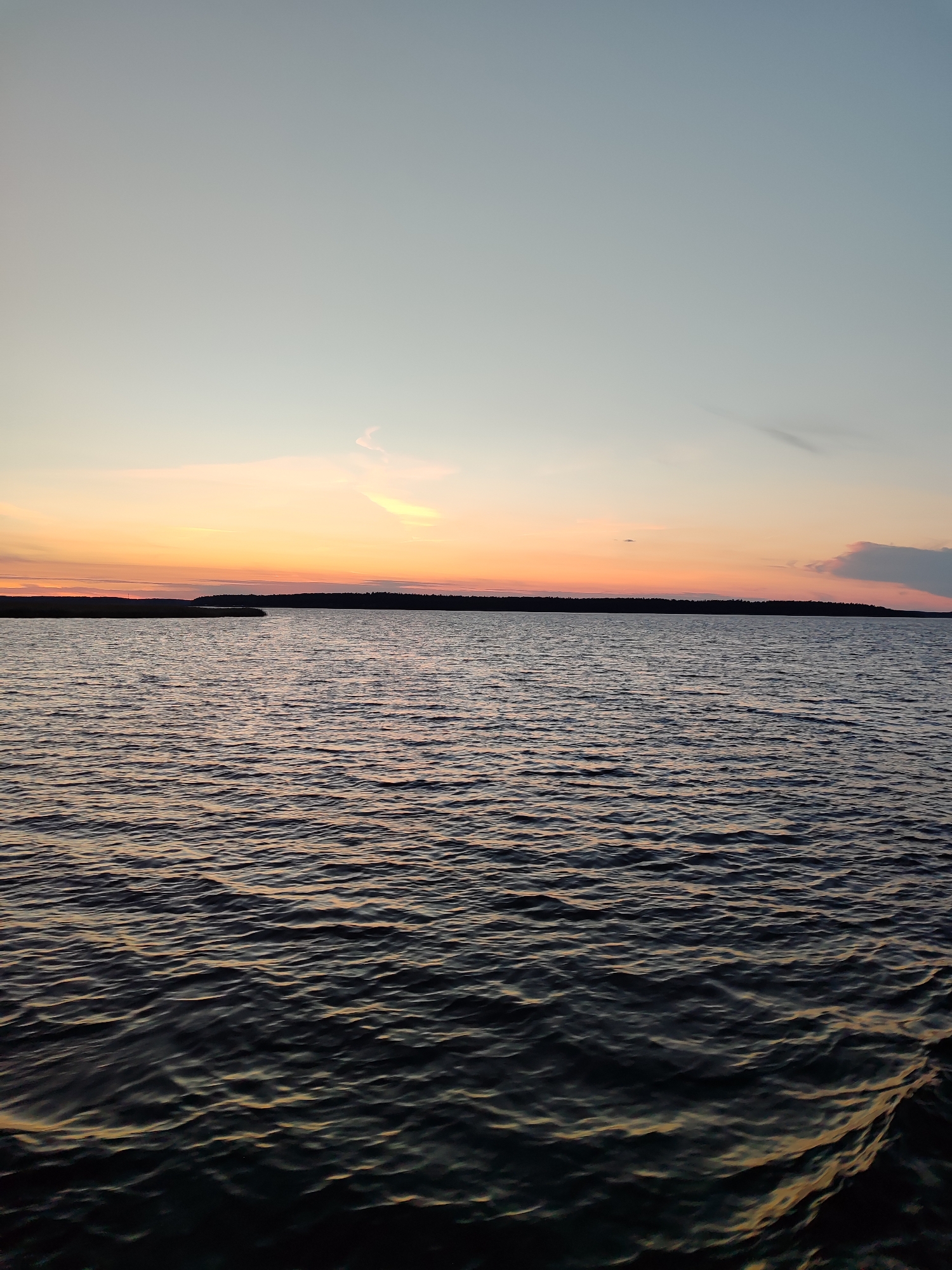 A body of water is shown during sunset. The sky is a gradient of light blue at the top, transitioning to orange and pink hues near the horizon. A silhouetted treeline is visible in the distance, marking the edge of the land. The water is dark and rippled, reflecting the colors of the sunset in subtle streaks. The overall scene depicts a tranquil, natural landscape at dusk.