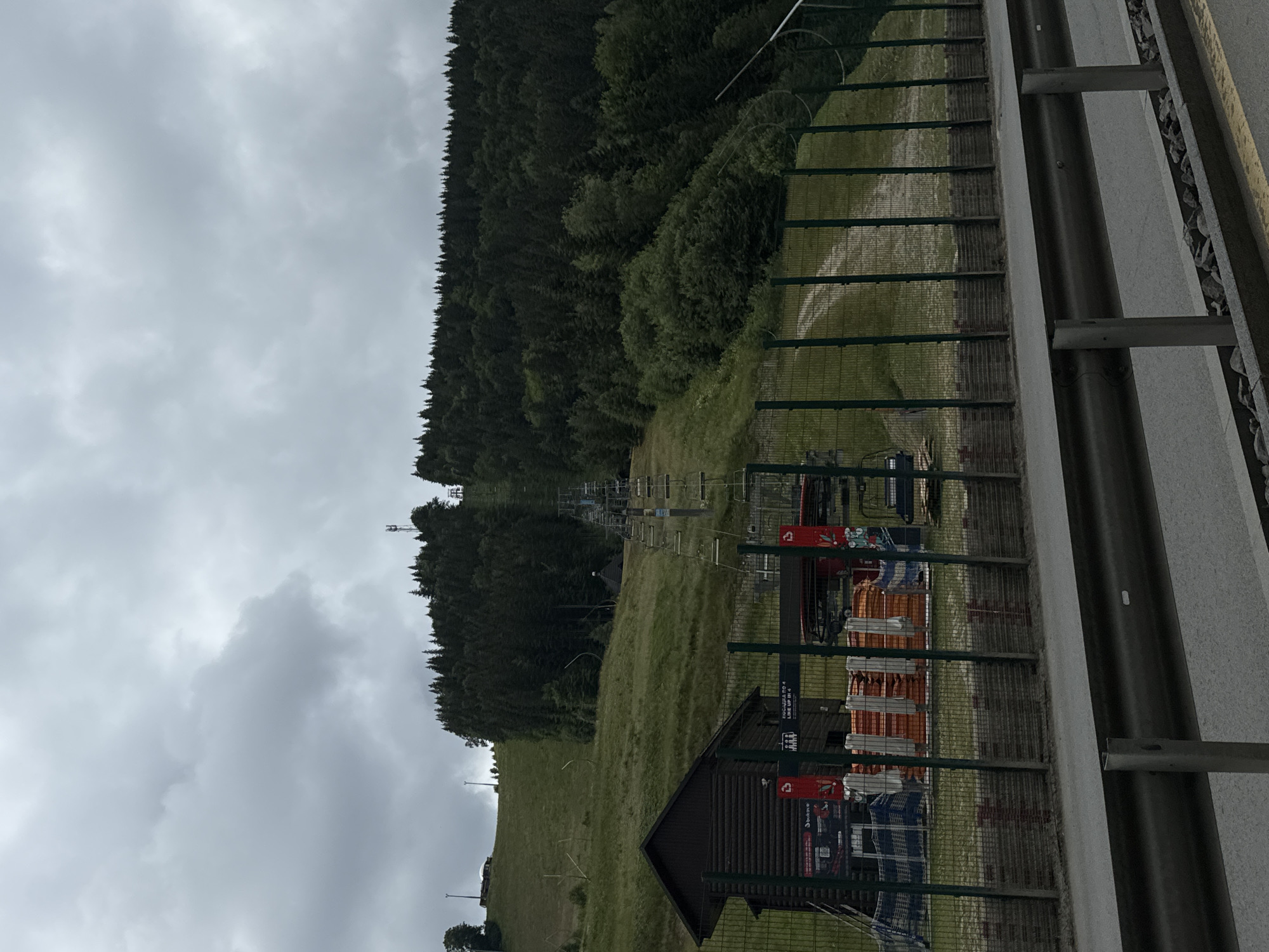 This is a full-frame, eye-level, outdoor, daytime photograph of a ski lift system on a cloudy day in Polianytsia, Ukraine. The ski lift stretches up a grassy, wooded mountainside. In the foreground, a metal guardrail separates a paved road from a fenced-off area. Within the fenced area, a small, dark wooden building stands to the left. Behind the building, stacks of orange and white objects, possibly safety equipment or snow-making materials, are visible, along with a ski lift loading station. The ski lift chairs are suspended on a cable that ascends the mountain. The mountain itself is covered in green grass and dark evergreen trees. A dirt path or track is visible winding up the mountainside behind the fence. The sky is overcast with thick grey clouds.
