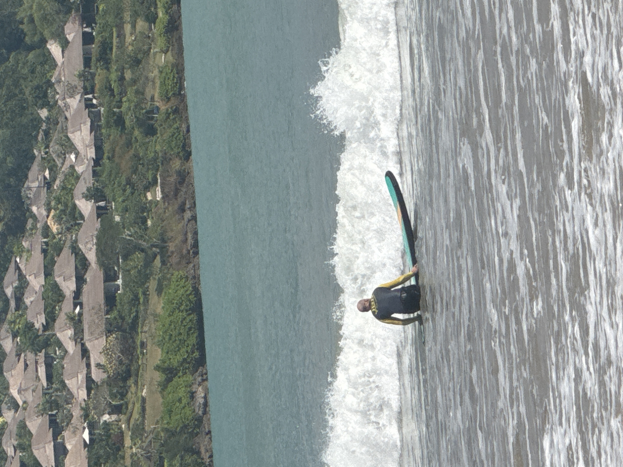 An overhead, slightly angled shot captures a surfer in shallow ocean water off a tropical coastline. The surfer is lying prone on a dark surfboard, which has a distinct light blue or green stripe. They are wearing a wetsuit that is black with bright yellow or green sleeves, and appear to be paddling or waiting for a wave.
To the left of the surfer, a white wave is breaking, indicating the surf line. The water transitions from a calm, deeper teal further out to the frothy white of the breaking wave, and then to a lighter, rippled grey-blue closer to the shore.
The background reveals a verdant coastline with dense green vegetation, primarily trees and bushes. Numerous buildings with pitched, grey-tiled roofs are visible, nestled among the greenery. These structures suggest a resort or village setting.
The scene is brightly lit, indicating it is daytime with clear weather. The overall impression is that of a calm day at a tropical beach, with the surfer engaging in their activity. Given the location context of Kuta Selatan, Indonesia, the architectural style and lush foliage are consistent with the region. There is no visible text in the image.