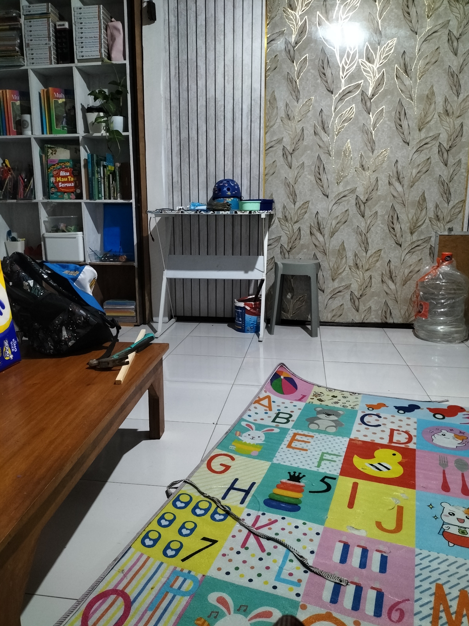 The image captures an indoor scene, likely a living room or playroom, with a mix of organizational furniture and recreational items. A tall, white bookshelf unit is visible on the left, filled with books and other items. In the center, a white desk with a striped blind behind it holds a blue cap and some other objects. To the right, a wall is covered with patterned wallpaper featuring stylized leaves in muted tones. In the foreground, a wooden table is positioned on the left, with a black bag and a hammer lying on it. A colorful alphabet mat with various cartoon images is spread out on the tiled floor in front of the desk. A large water dispenser is situated to the right of the patterned wall. The overall impression is of a domestic space that serves both functional and recreational purposes. No specific landmarks or indicators point to the city of Malang, Indonesia.