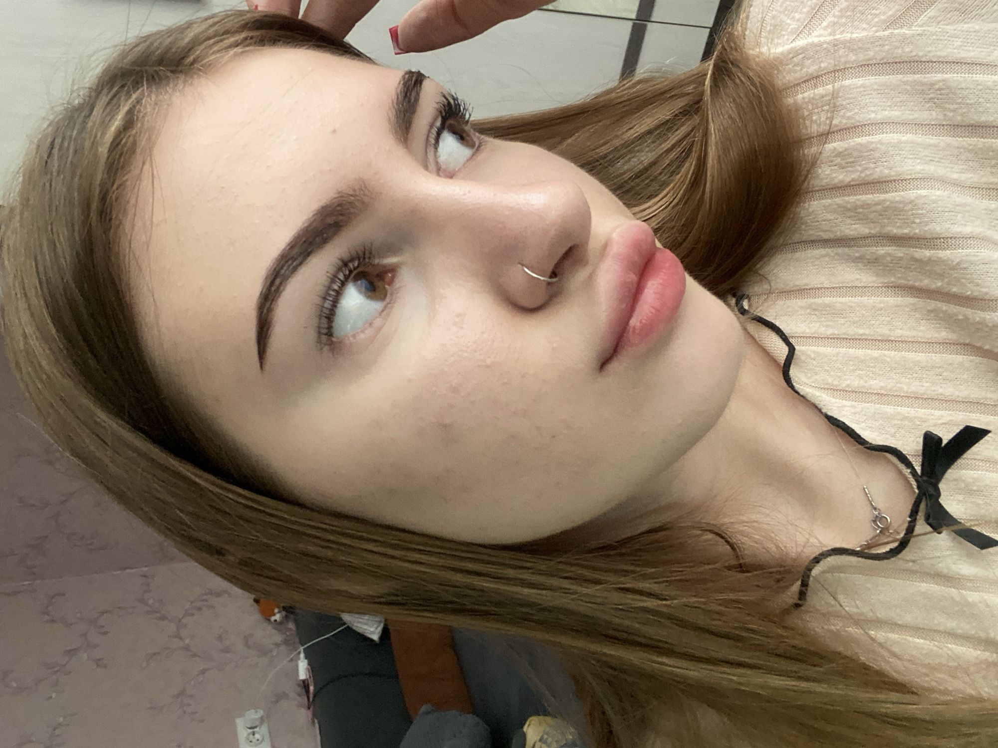 The image shows a close-up, profile view of a young woman with light brown hair, looking to her right. She is wearing a light-colored, ribbed sweater with black trim and a black bow detail. A silver necklace with a small pendant is visible around her neck. She has a silver hoop nose ring and appears to be touching her forehead with her right hand. The background is softly blurred, showing a wall with patterned wallpaper, an electrical outlet, and a glimpse of furniture. The scene is indoors, likely in a residential setting within the city of Dyakovo, Russia.